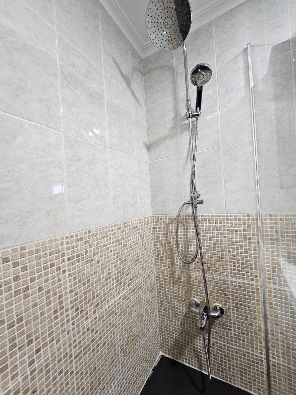 Ducha contemporánea con sistema de doble rociador, combinando azulejos de efecto mármol y mosaico en tonos tierra.