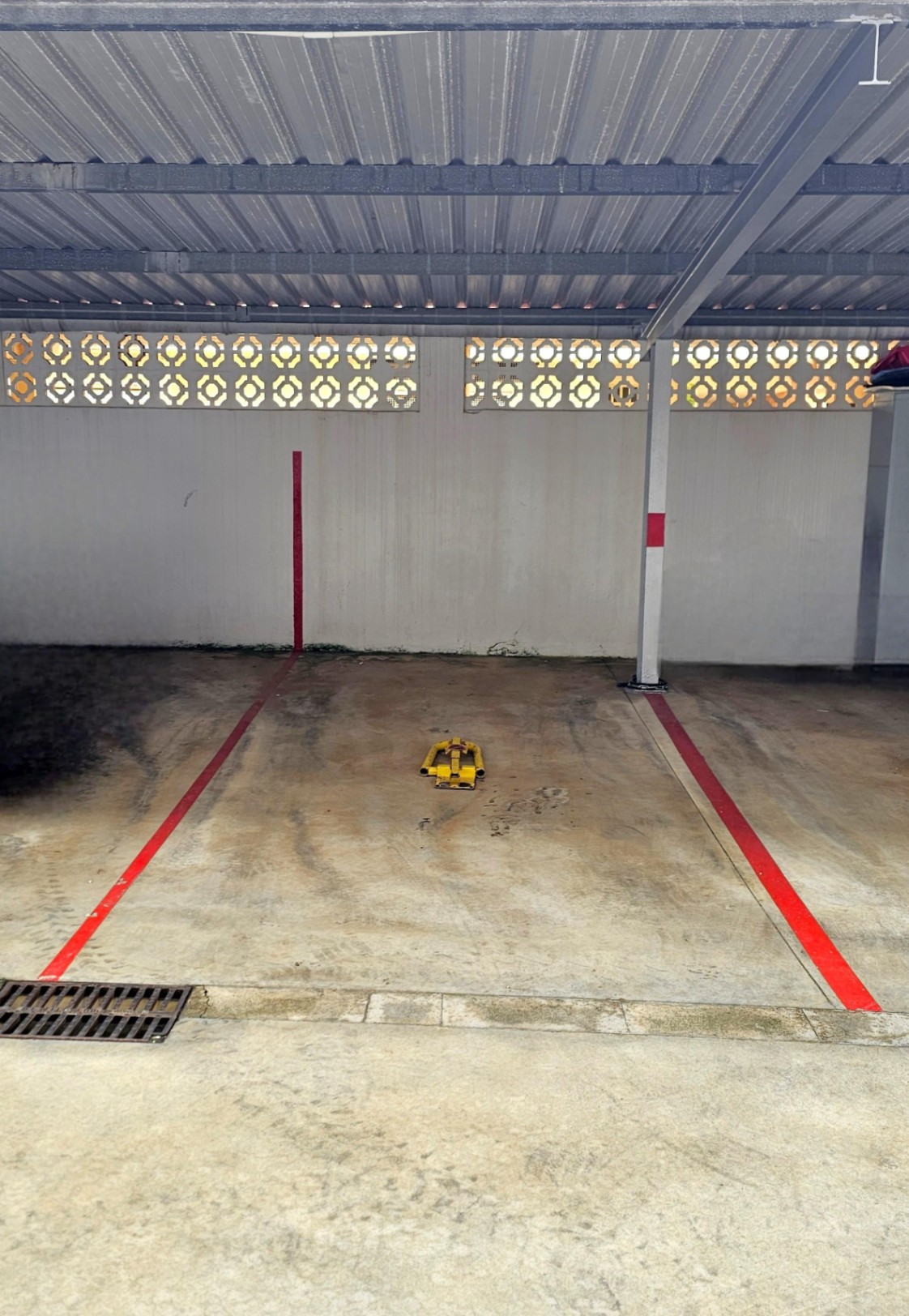 Plaza de parking privada y cubierta con techado metálico, delimitada por líneas rojas y equipada con un cepo de seguridad amarillo sobre suelo de hormigón.