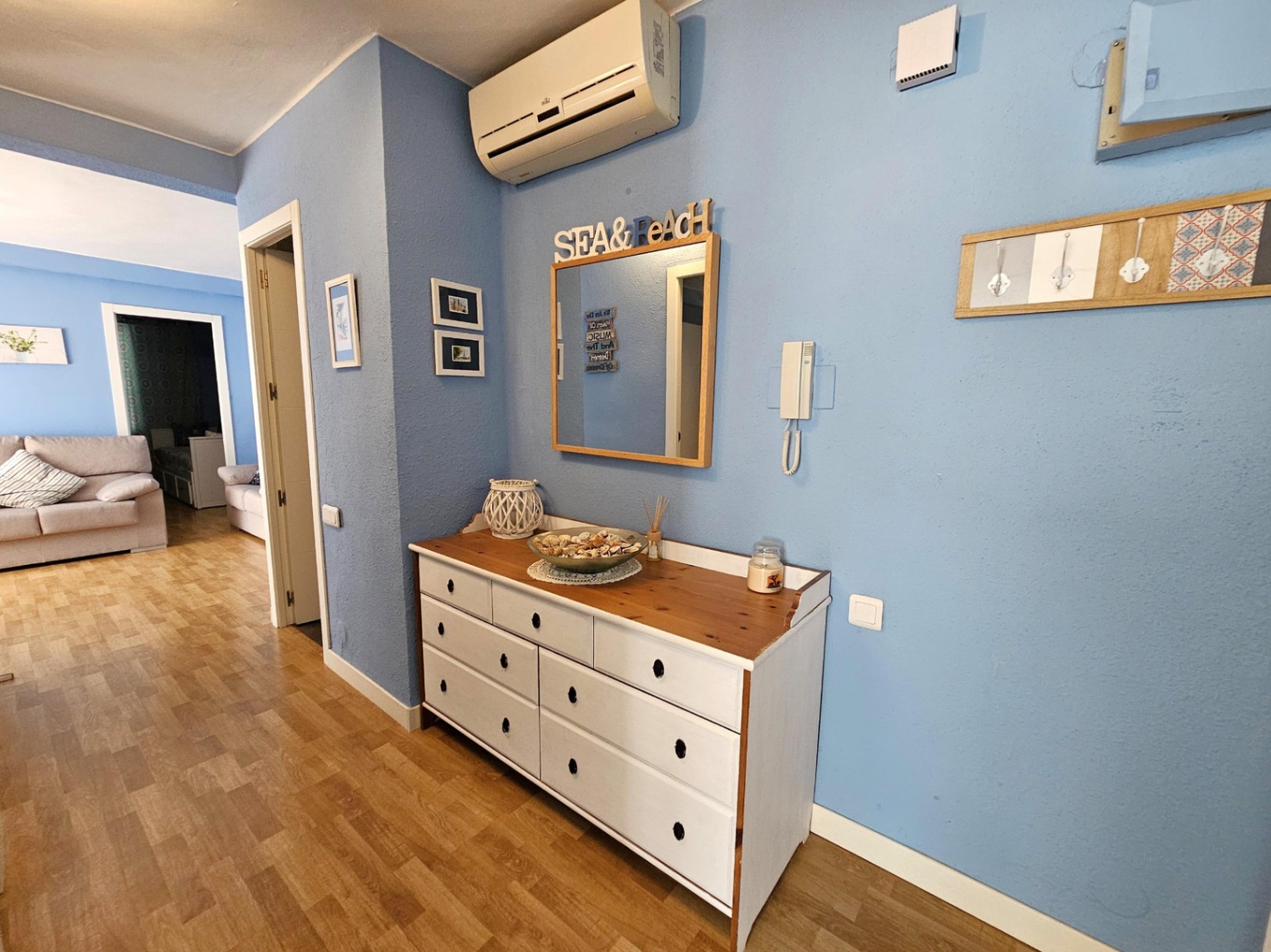 Recibidor con paredes azules, cómoda de madera blanca, suelos de parqué y aire acondicionado split, con vista directa hacia el salón.