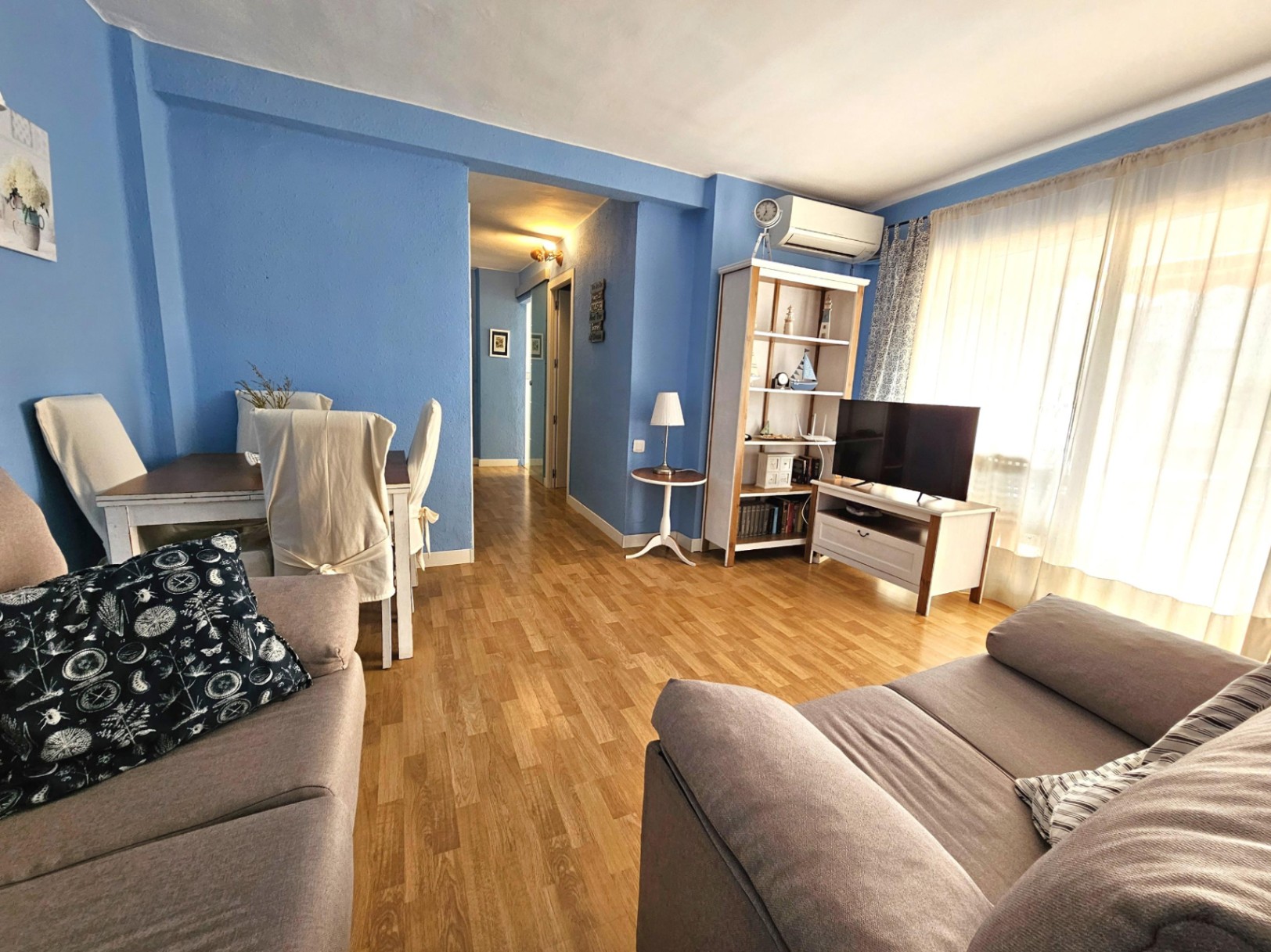 Salón comedor con paredes azules, suelos de parqué y aire acondicionado. Incluye zona de estar con sofás y acceso directo a un pasillo distribuidor.