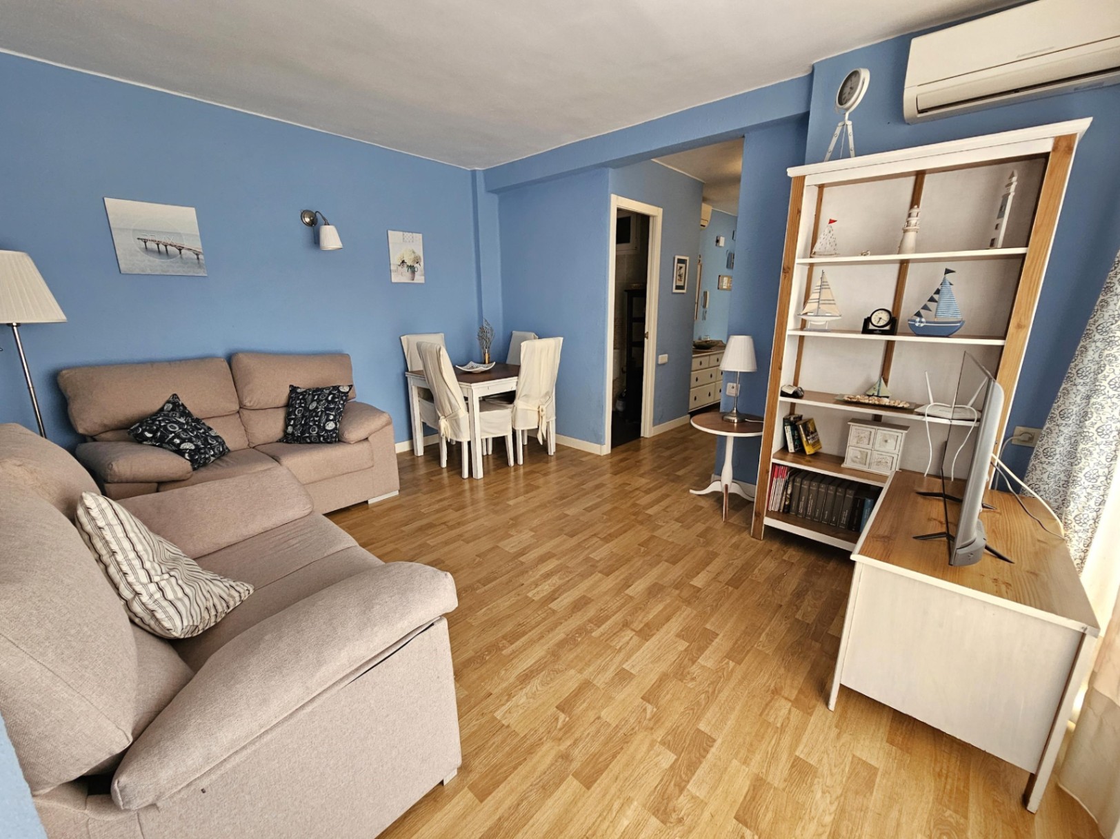 Salón comedor con paredes en tono azul, suelos de parqué y mobiliario contemporáneo. Dispone de aire acondicionado split y una estantería de madera con motivos náuticos.