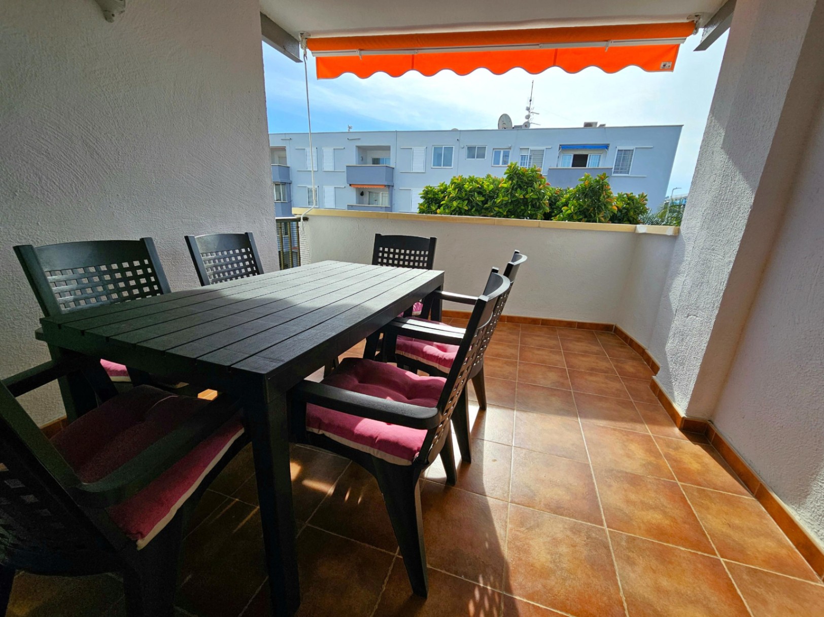 Terraza exterior con suelo cerámico, mesa de comedor negra para seis comensales, cojines rosados y toldo extensible naranja con vistas despejadas.