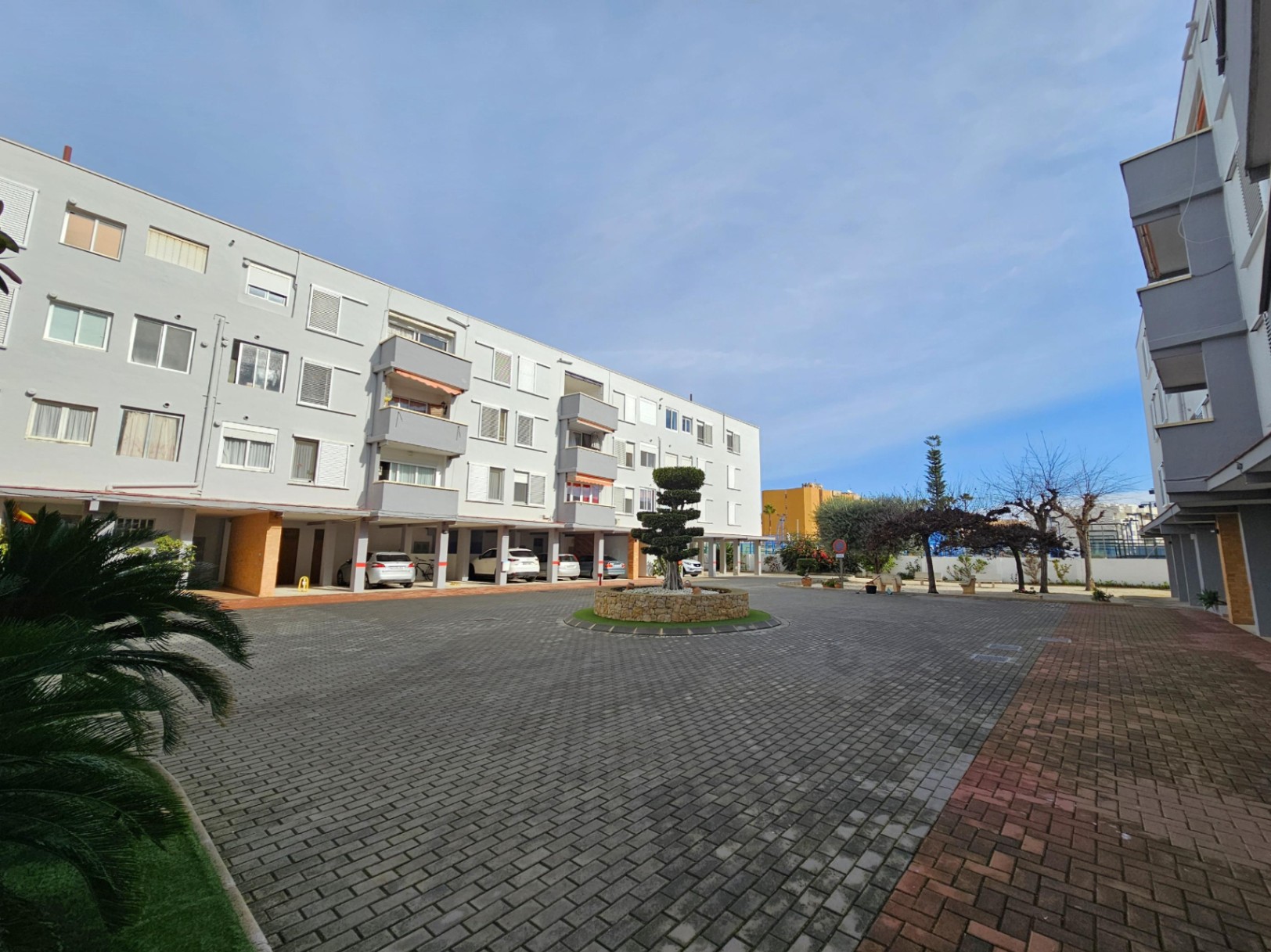 Vista exterior del complejo con fachada gris, zona de aparcamiento cubierto y patio pavimentado con una jardinera central de piedra.