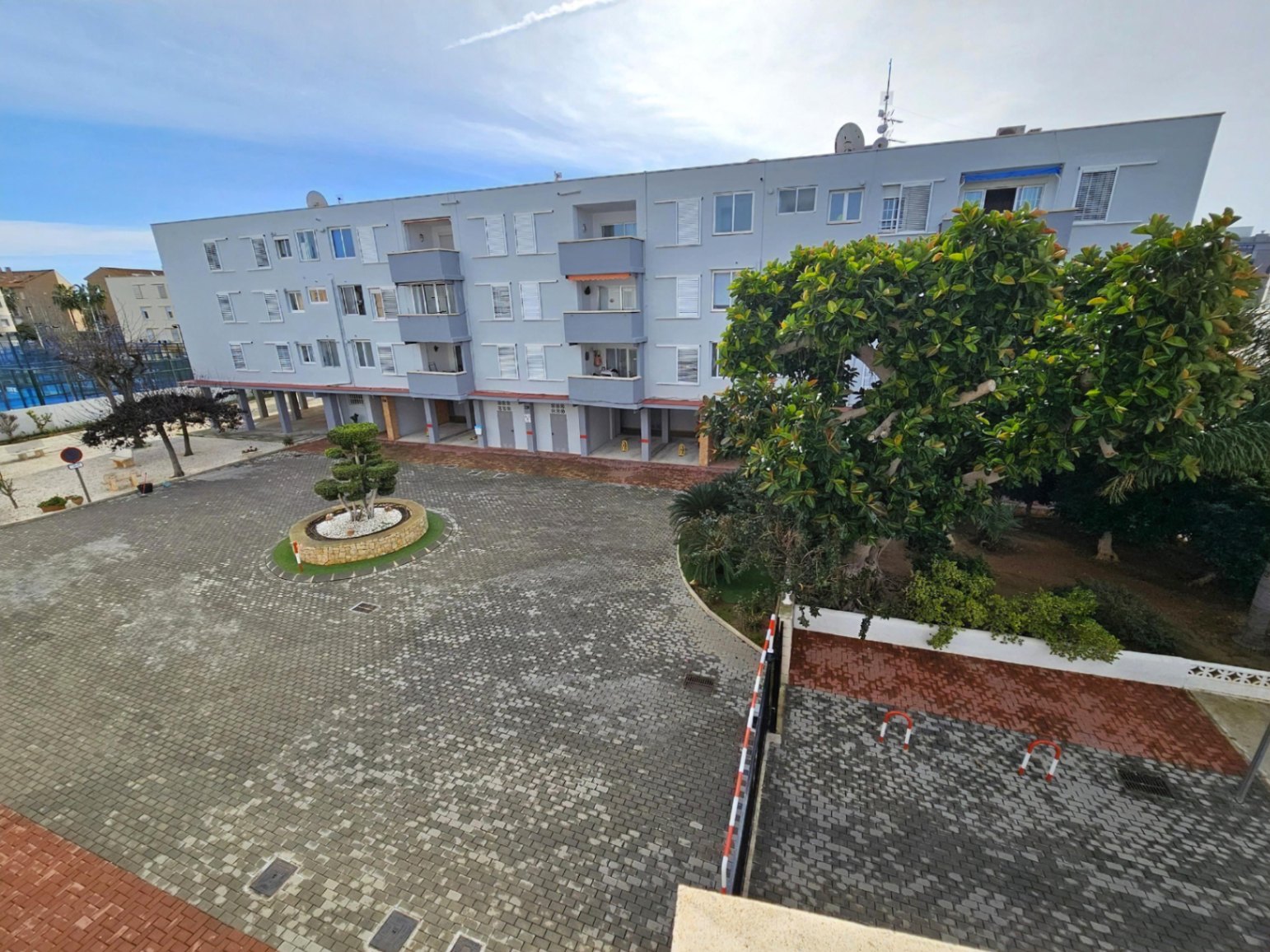 Vista exterior del complejo que muestra el patio pavimentado, zonas ajardinadas y el acceso al parking cubierto privado bajo el edificio residencial.