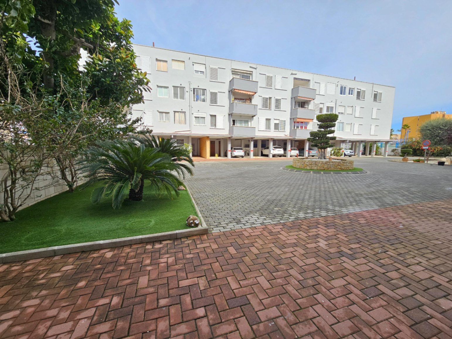 Vista exterior del edificio residencial con zona de aparcamiento cubierto, jardines cuidados y pavimentación de adoquines en el Arenal de Jávea.
