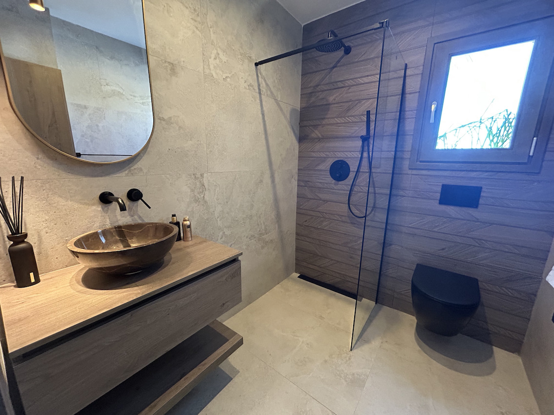 Baño moderno con ducha italiana, grifería negra mate, lavabo de piedra sobre mueble flotante de madera y revestimientos cerámicos texturizados.
