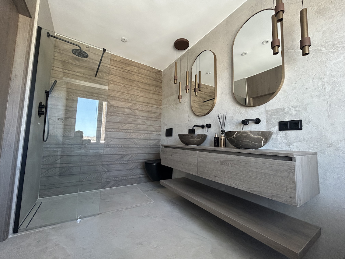 Baño moderno con lavabo doble de piedra, grifería negra empotrada, espejos ovalados y ducha de obra con revestimiento cerámico efecto madera.