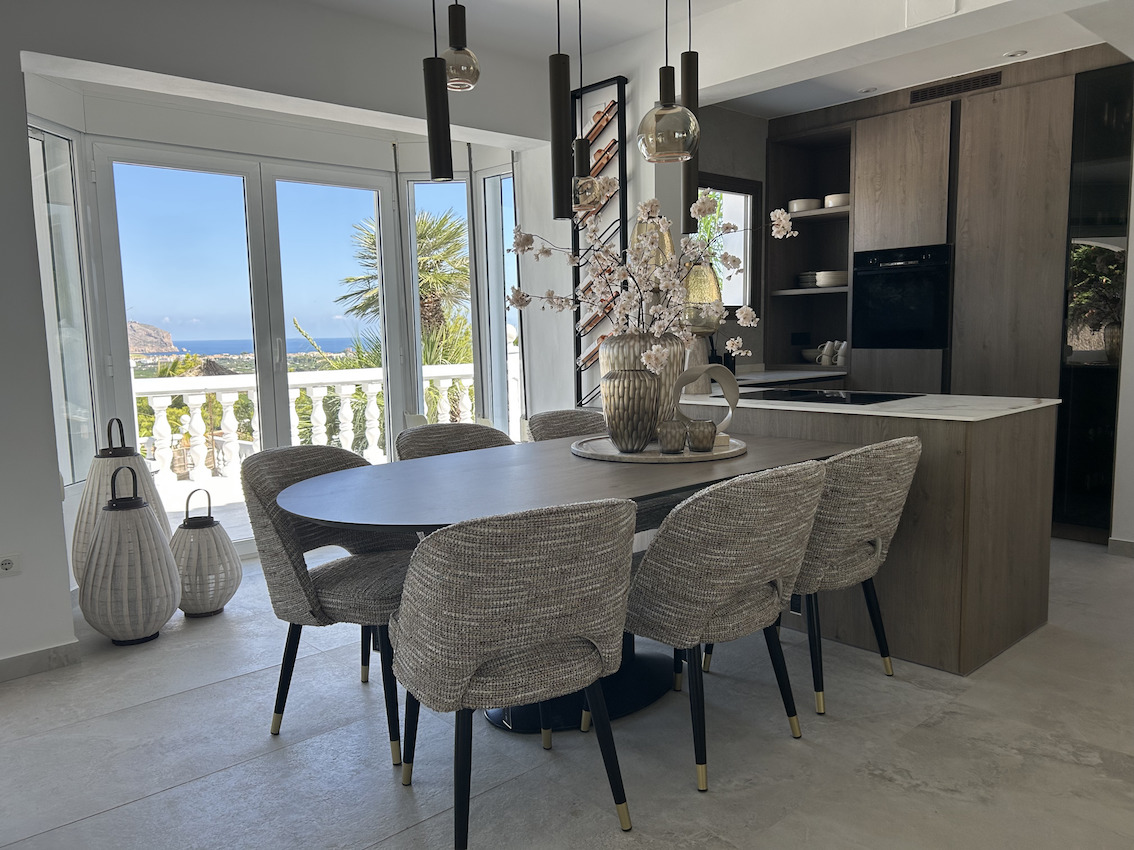 Comedor abierto con mesa ovalada y sillas de diseño junto a una cocina moderna. Los grandes ventanales ofrecen vistas despejadas al mar y acceso a la terraza.