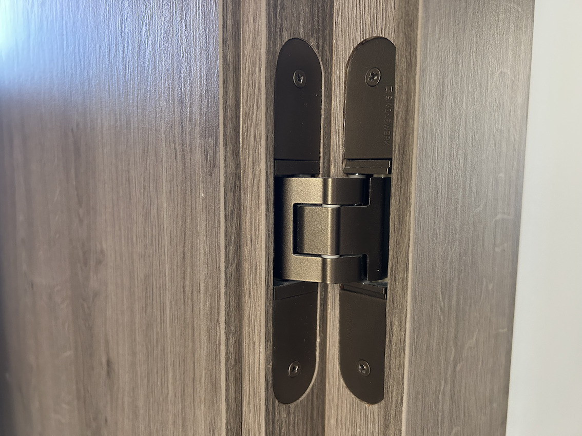 Detalle de bisagra oculta de alta calidad en acabado bronce, integrada en carpintería de madera con textura natural, reflejando los acabados premium de la reforma de 2025.