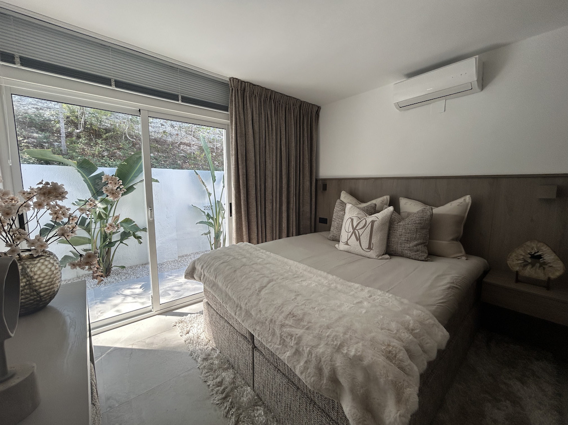 Dormitorio moderno con acceso directo al patio mediante ventanal corredero, aire acondicionado y cabecero de madera con iluminación integrada.