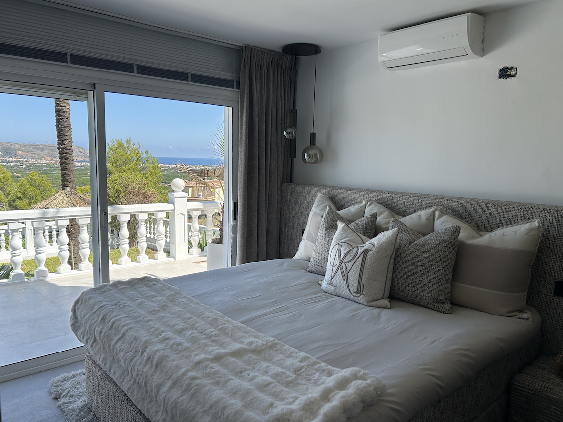 Dormitorio principal con cabecero tapizado, aire acondicionado y acceso directo a una terraza privada con vistas panorámicas al mar y al entorno natural.