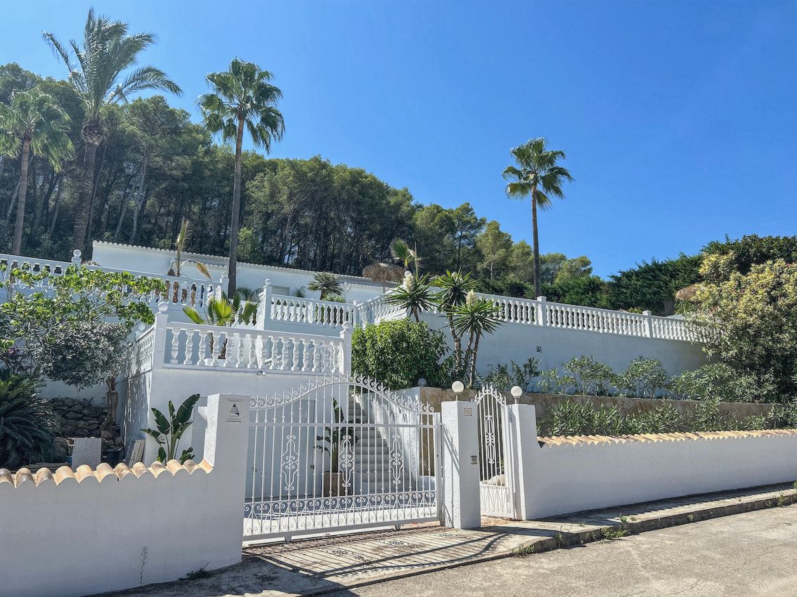Fachada escalonada con balaustradas blancas, portón de hierro forjado y jardines mediterráneos con palmeras bajo un cielo despejado.