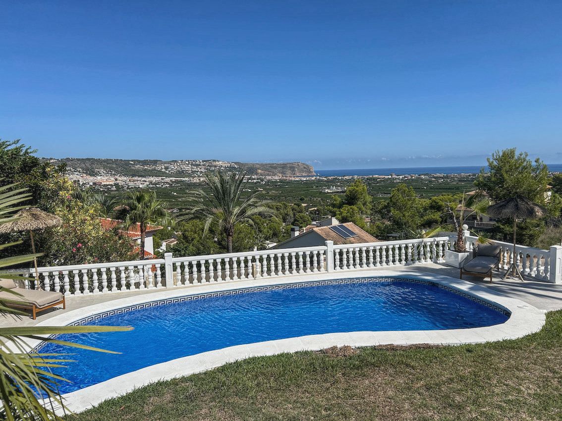 Piscina de diseño curvo rodeada por una balaustrada blanca, con vistas panorámicas al valle y al mar Mediterráneo bajo un cielo despejado.