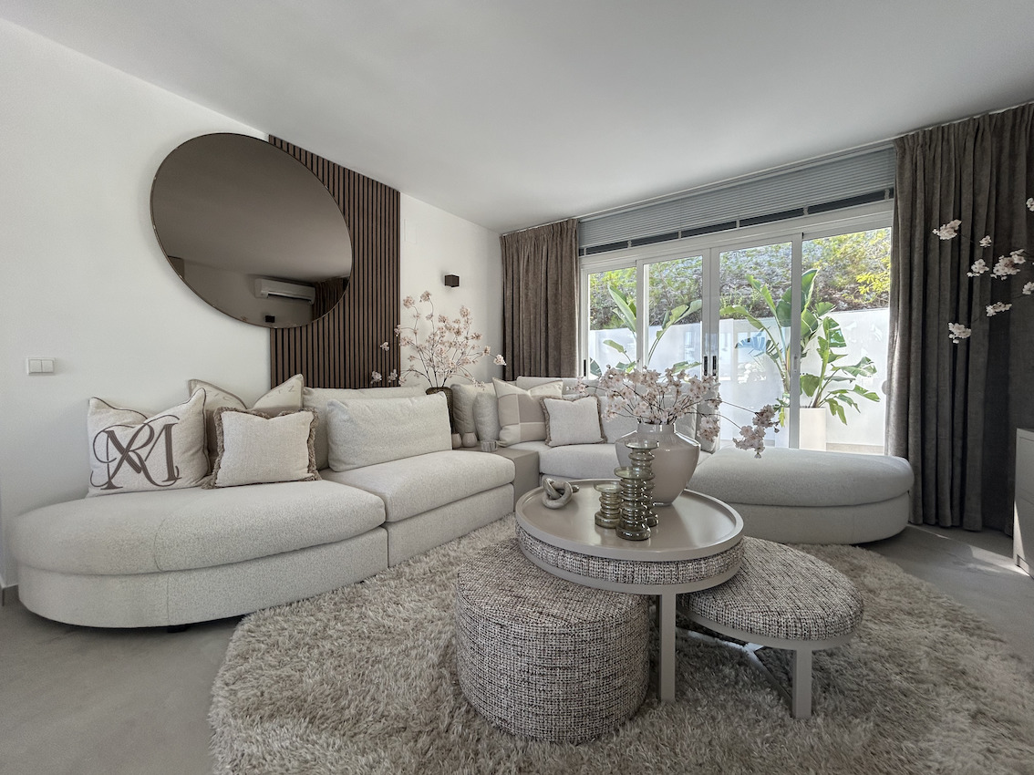 Salón moderno con sofá modular curvo, pared decorativa de listones de madera con espejo circular y amplios ventanales que conectan con la terraza exterior.