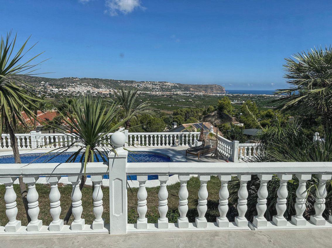 Terraza elevada con balaustrada blanca clásica que ofrece vistas panorámicas al valle, la costa y la piscina privada rodeada de vegetación mediterránea.