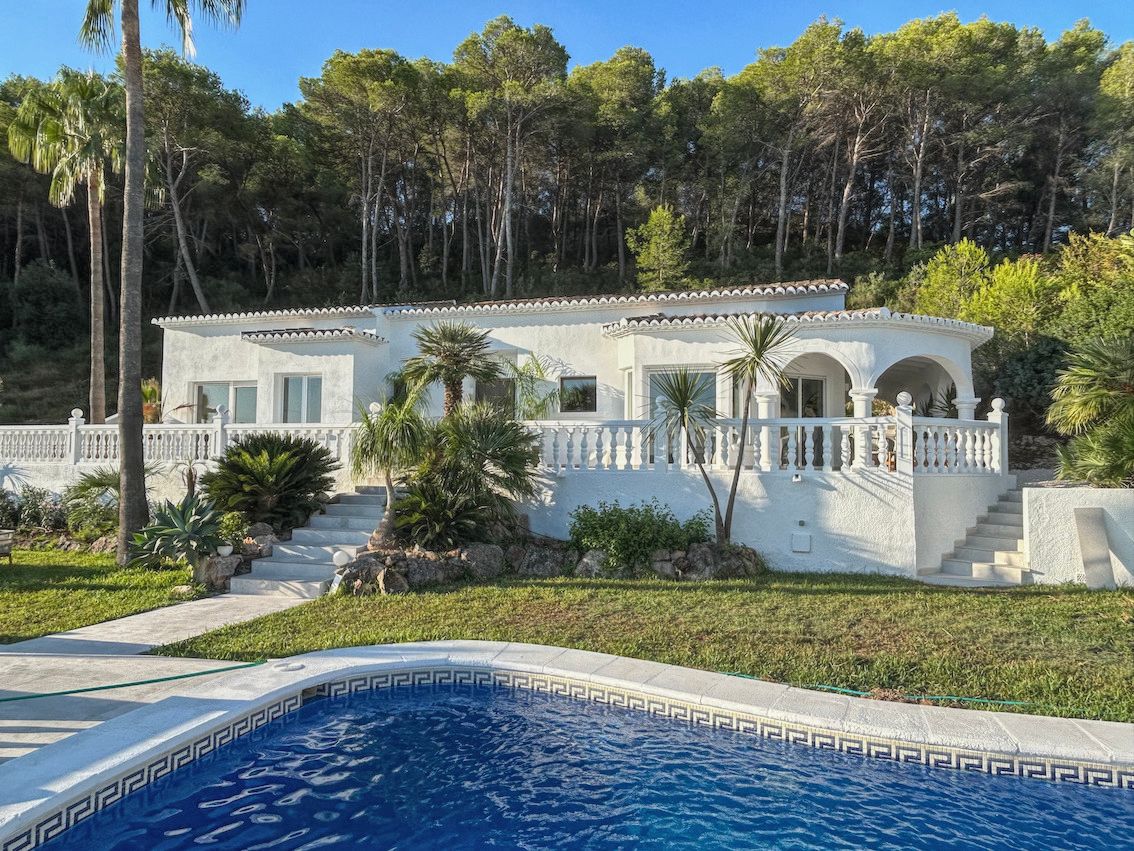 Villa blanca de estilo mediterráneo con amplia terraza de balaustrada, piscina con cenefa clásica y entorno natural rodeado de pinos.