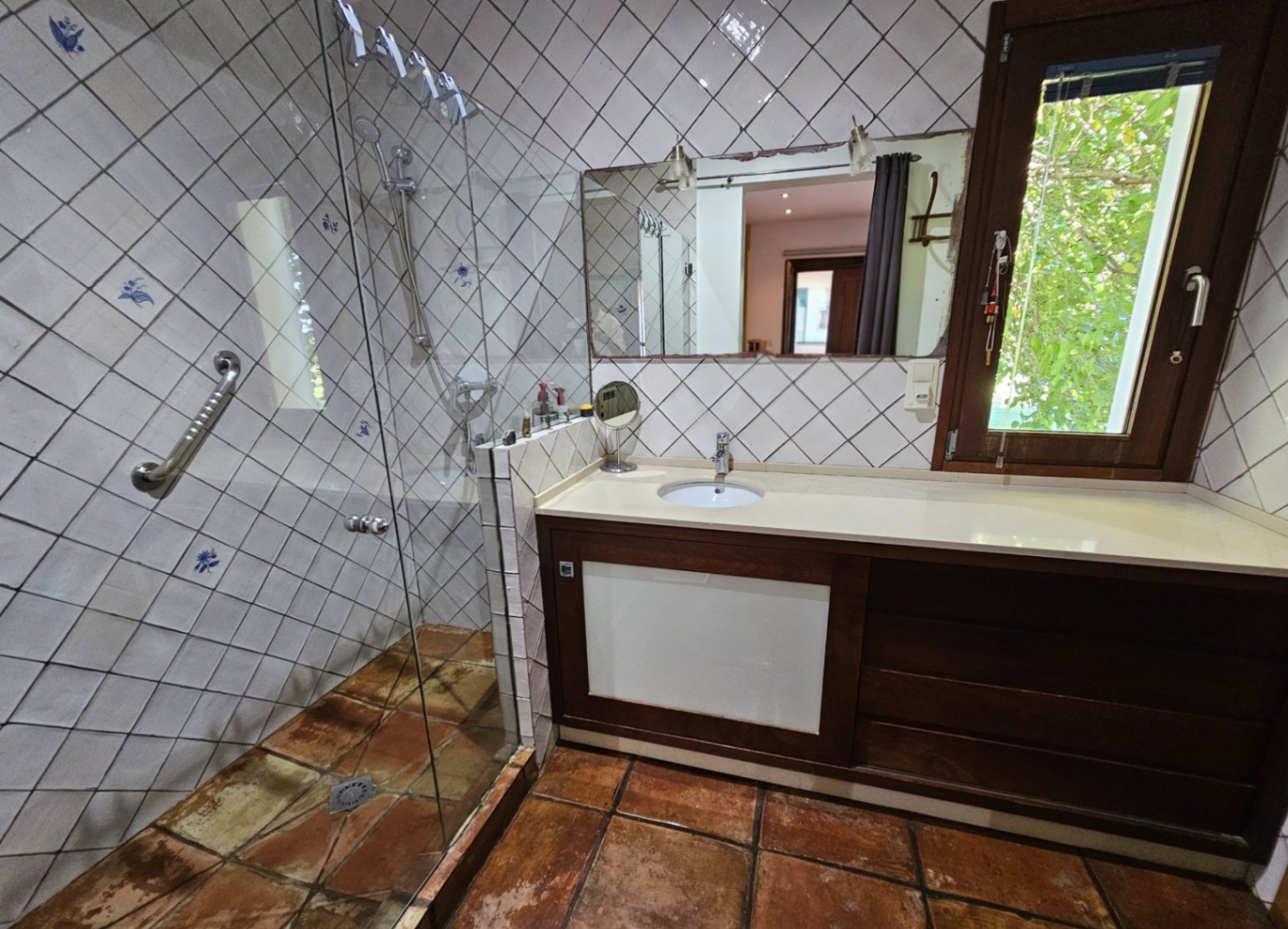 Baño con ducha acristalada, azulejos artesanales pintados a mano y suelos de barro. Incluye un mueble de madera oscura y ventana con vistas al jardín.