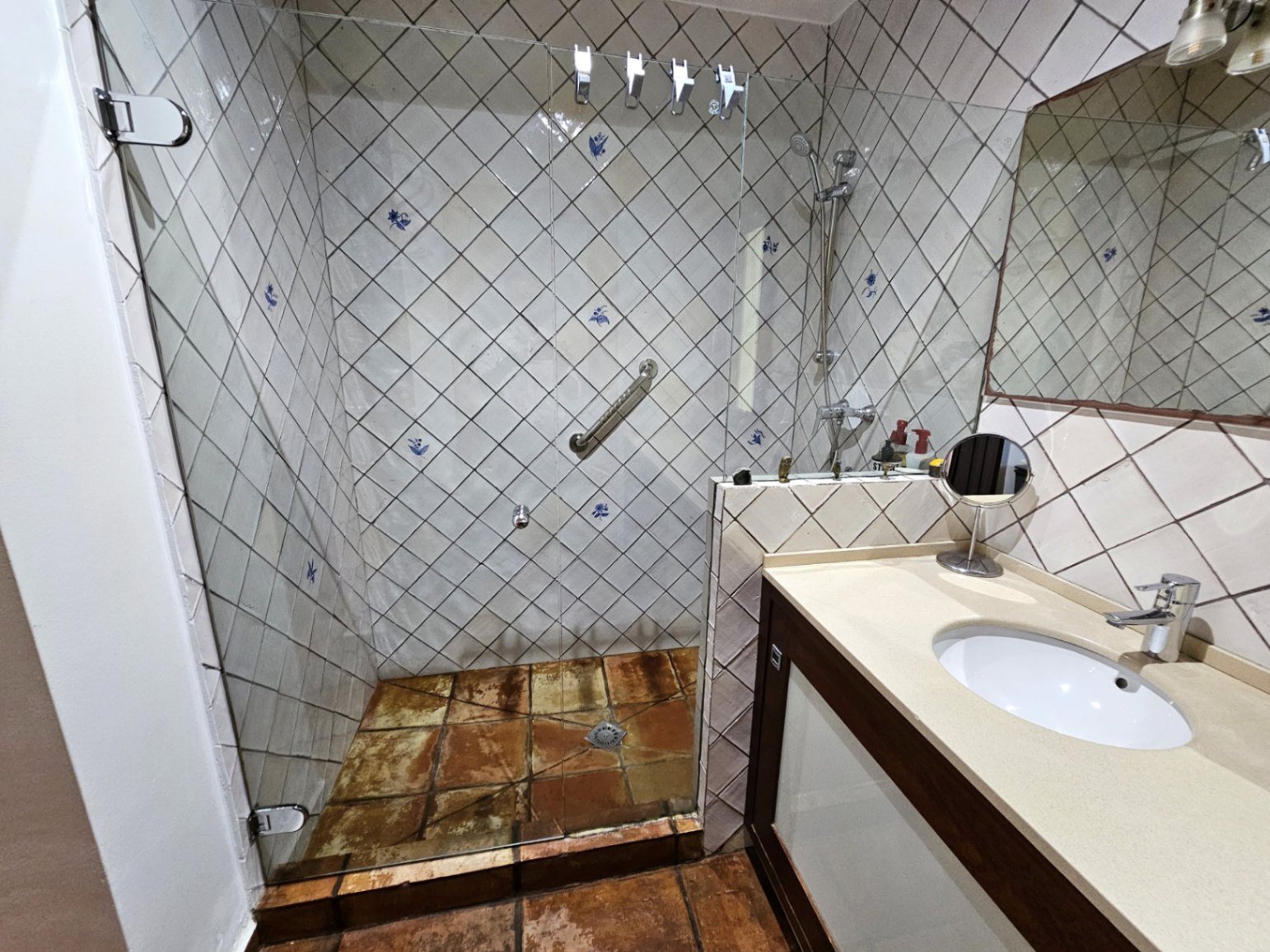 Baño con ducha de obra, mampara de cristal y azulejos artesanales pintados a mano. Destacan los suelos de barro cocido y el mueble de lavabo con encimera en tonos crema.