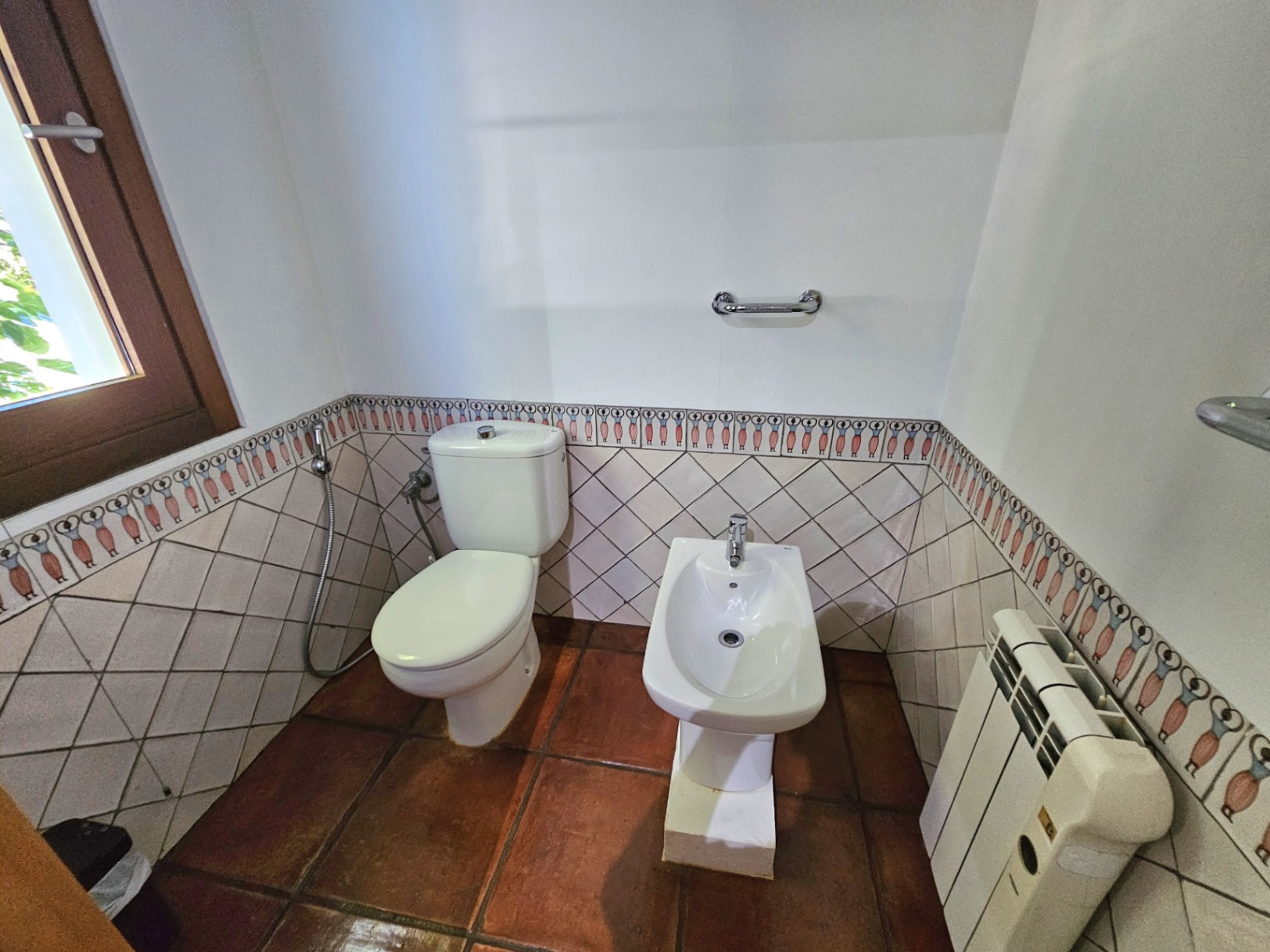 Baño con inodoro y bidé, destacando el suelo de barro artesanal y el alicatado con cenefa pintada a mano. Incluye radiador y ventana con marco de madera.