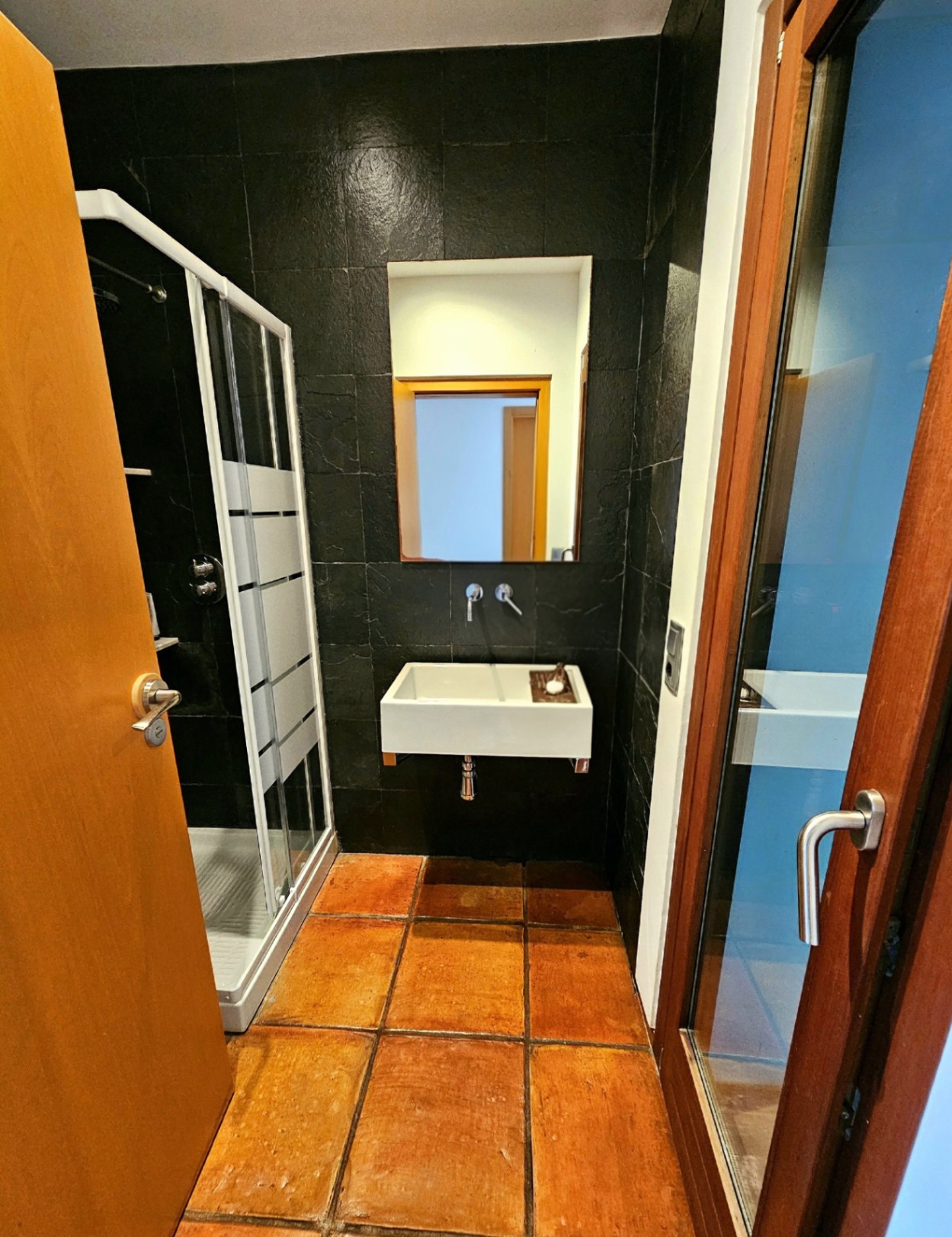 Baño con suelos de barro artesanal, paredes de azulejo oscuro texturizado, lavabo suspendido de diseño moderno y cabina de ducha con perfiles blancos.