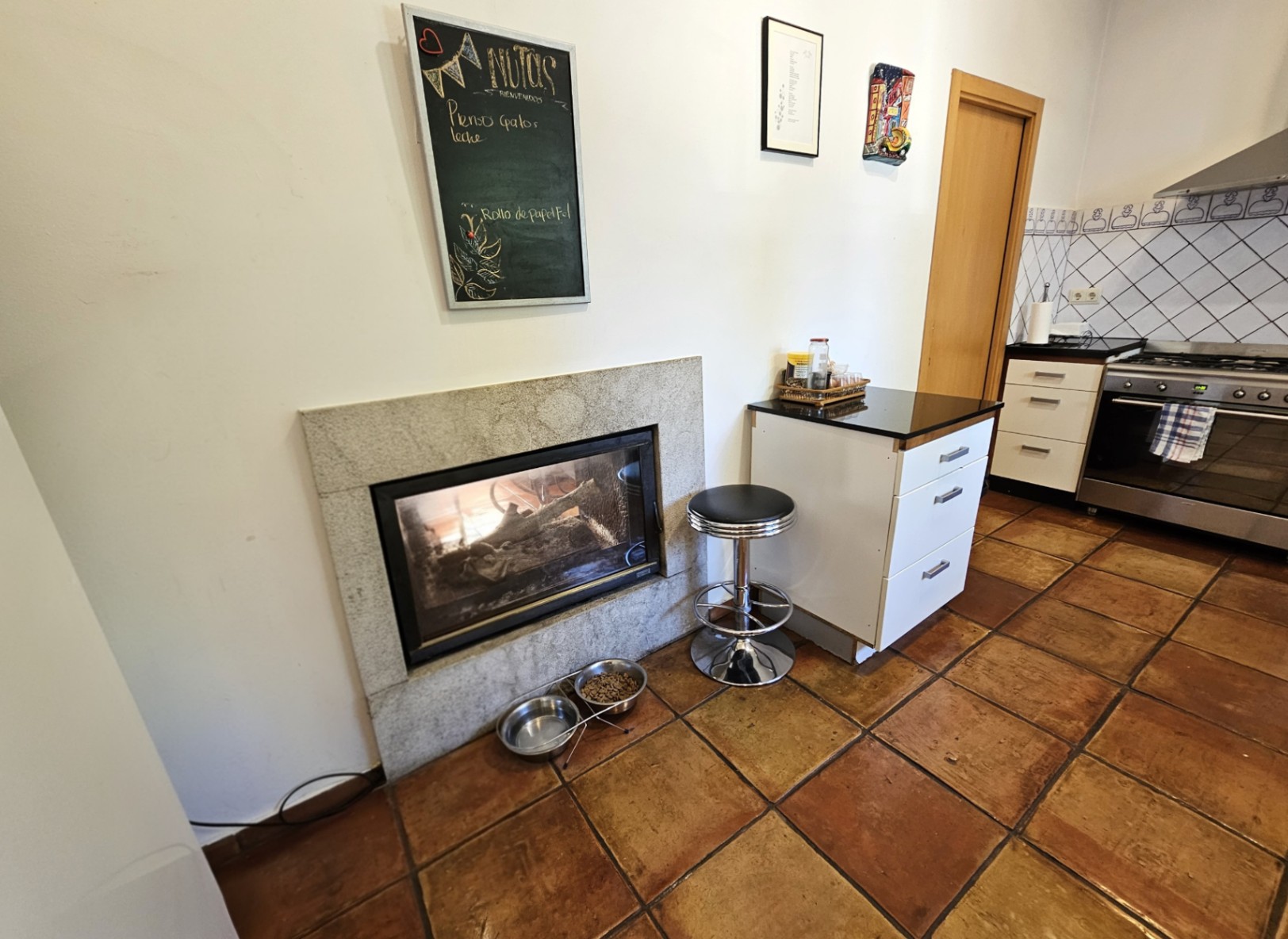 Cocina con suelos de barro artesanal y chimenea encastrada con marco de piedra. Incluye mobiliario blanco, encimera oscura y azulejos decorativos pintados a mano.