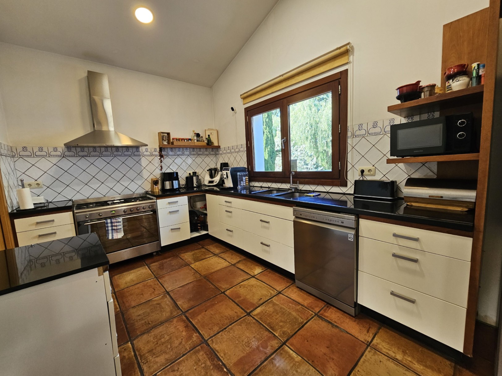 Cocina equipada con suelos de barro hechos a mano, encimeras oscuras y electrodomésticos de acero inoxidable, incluyendo una amplia cocina de gas y campana extractora.