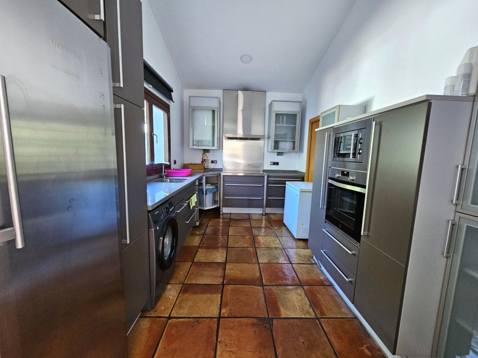 Cocina moderna con armarios en tono gris, electrodomésticos de acero inoxidable y suelos de barro hechos a mano. Incluye lavadora integrada y encimeras blancas.