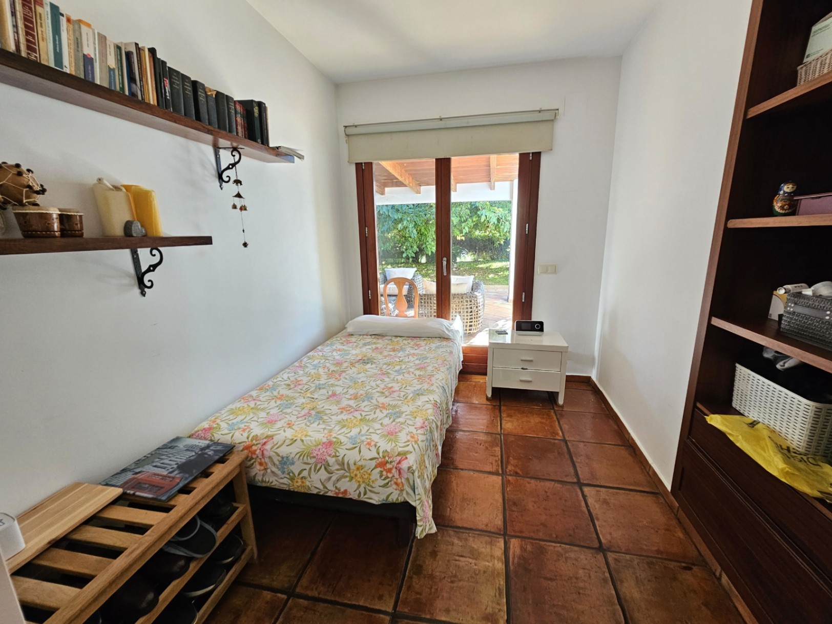 Dormitorio individual con suelos de barro hechos a mano, estanterías de madera y acceso directo a la naya y el jardín a través de grandes ventanales.
