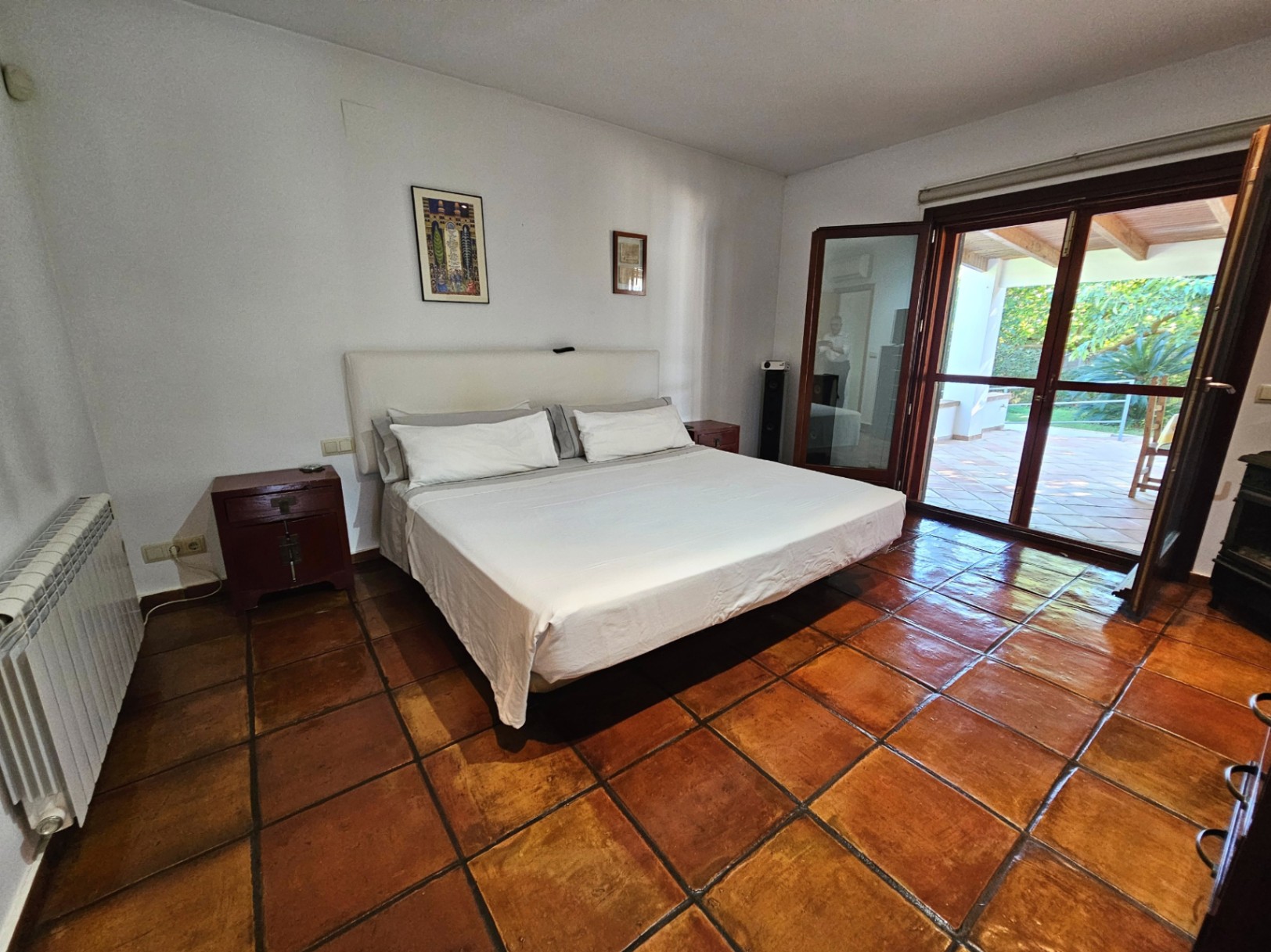 Dormitorio luminoso con suelos de barro hechos a mano, cama de matrimonio y acceso directo a la terraza exterior a través de grandes ventanales de madera.