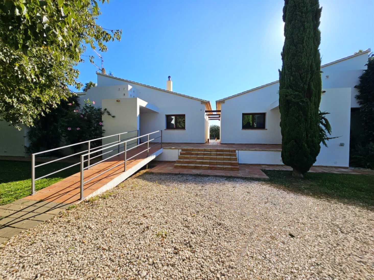 Fachada de la villa con dos alas independientes, rampa de acceso metálica, escalones de barro cocido y zona de aparcamiento de grava rodeada de cipreses.