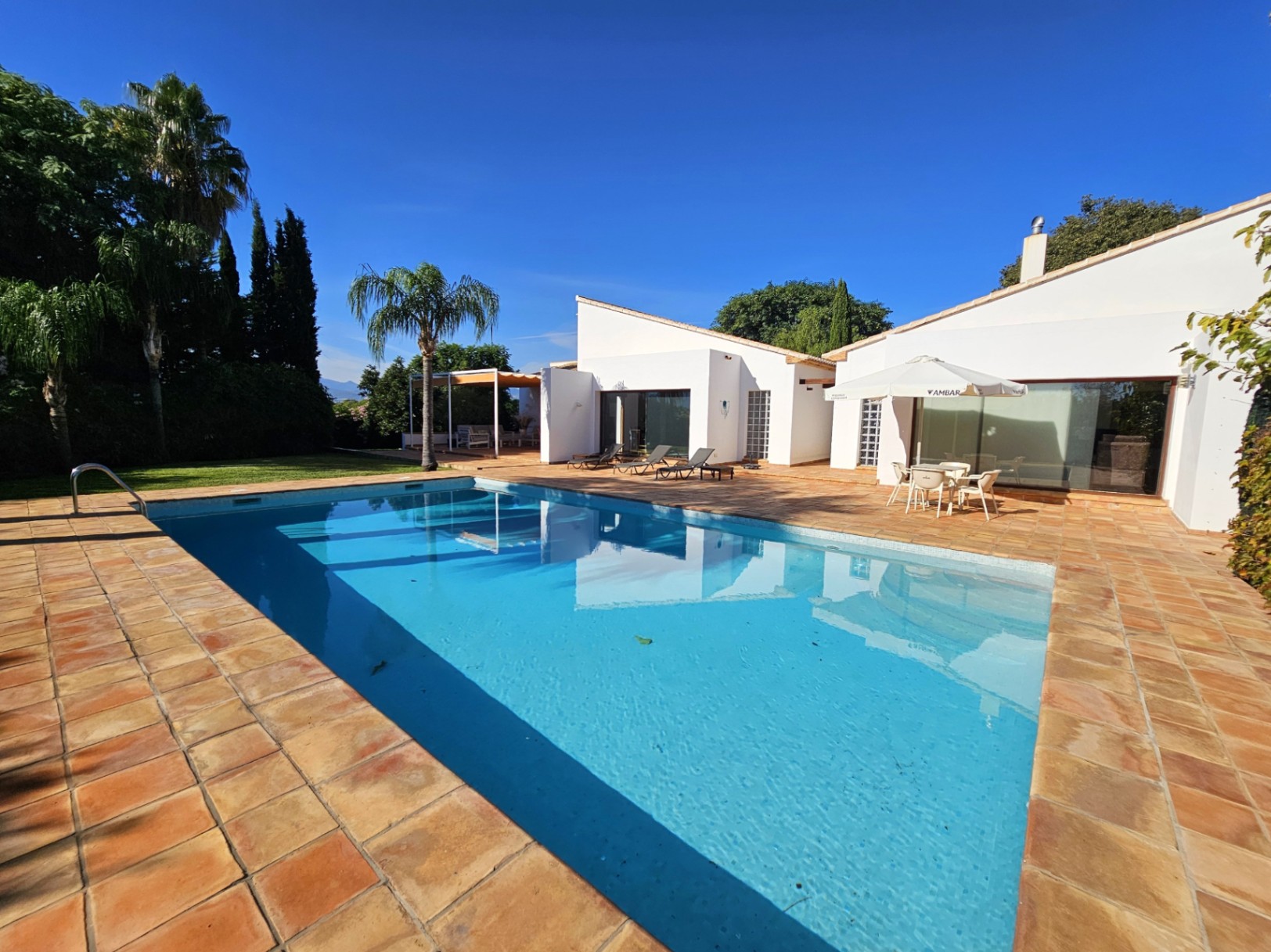 Piscina exterior rodeada de una amplia terraza de barro manual, con vistas a las fachadas blancas de la villa y zonas ajardinadas con palmeras bajo un cielo despejado.