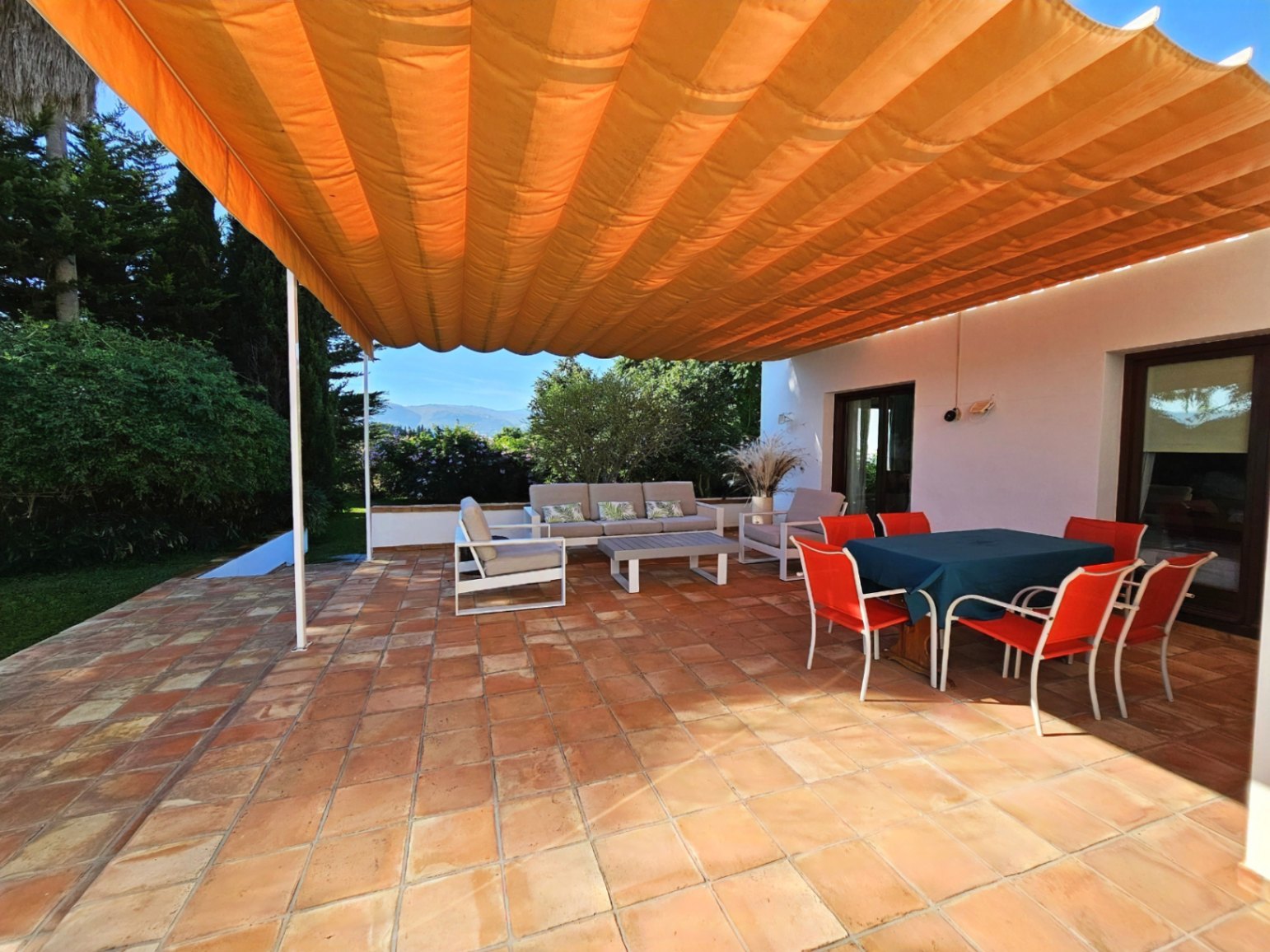 Terraza exterior con suelos de barro manual y un gran toldo naranja. Dispone de zona de comedor y estar con vistas despejadas al jardín y las montañas.