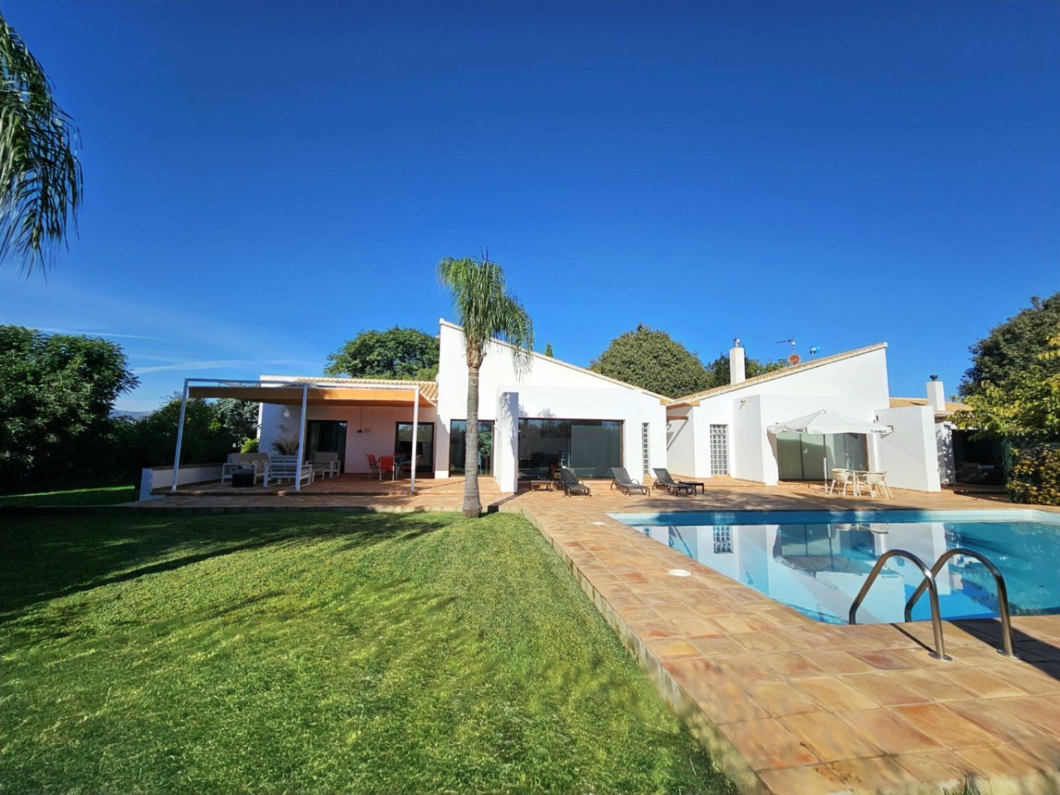 Vista exterior de la villa con piscina, rodeada de suelos de barro hechos a mano y una amplia zona de césped bajo el cielo despejado.