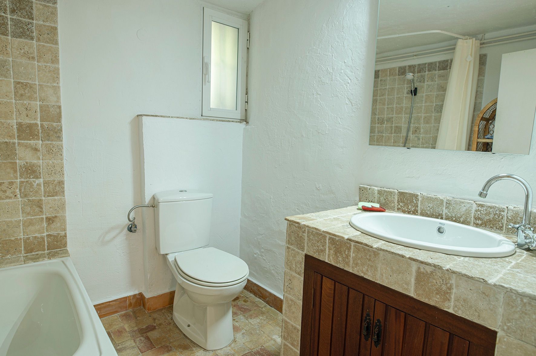 Baño de estilo rústico con encimera de piedra natural, mueble de madera oscura, lavabo ovalado y bañera blanca junto a una ventana de ventilación.