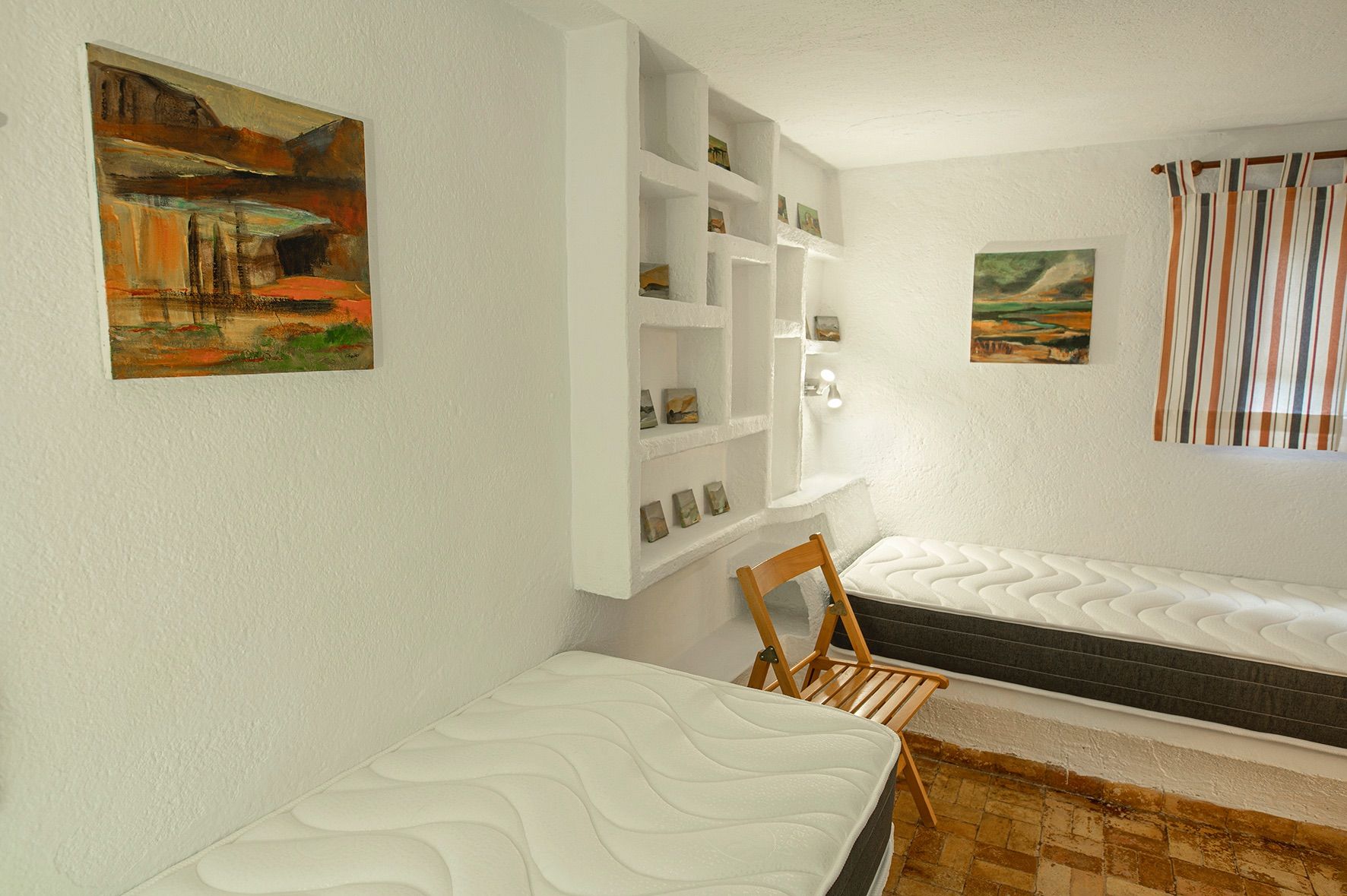 Dormitorio con dos camas individuales, paredes blancas de acabado rugoso y estantería de obra integrada. Suelo de baldosas de barro cocido y luz natural.