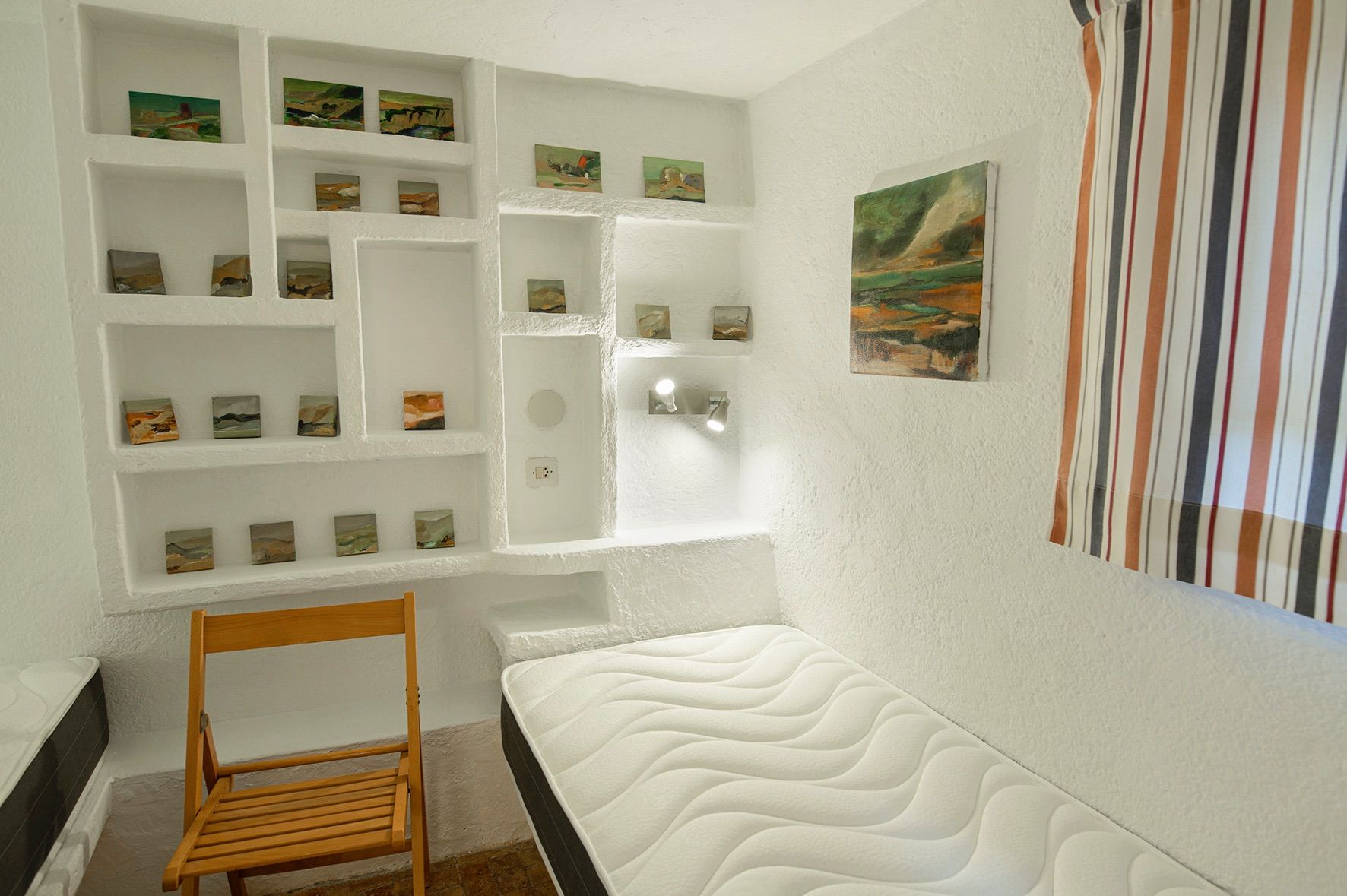 Dormitorio con estanterías de obra integradas en paredes blancas de textura rugosa, decoradas con pequeñas obras de arte y una silla plegable de madera.