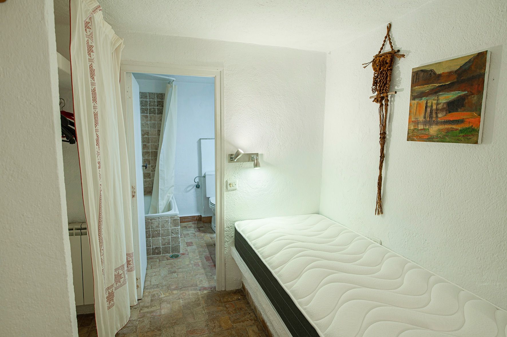 Dormitorio funcional con cama individual sobre base de obra, paredes blancas texturizadas y baño en suite con azulejos rústicos en tonos tierra.