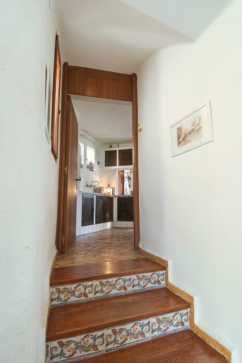Escalera con peldaños de madera y frontales de azulejo tradicional que conecta con una cocina rústica de armarios con celosía y suelos de barro.