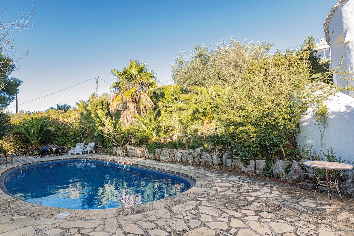 Piscina de forma curva rodeada de un solárium de piedra natural y vegetación mediterránea madura. Incluye zona de descanso con tumbonas y muros decorativos de piedra.