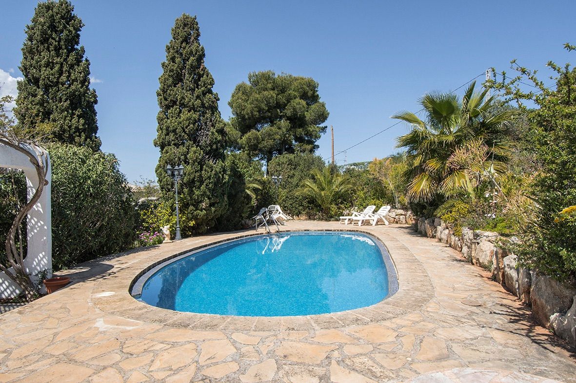 Piscina de forma ovalada rodeada por una terraza de piedra natural, jardines mediterráneos con cipreses altos y palmeras bajo un cielo despejado.