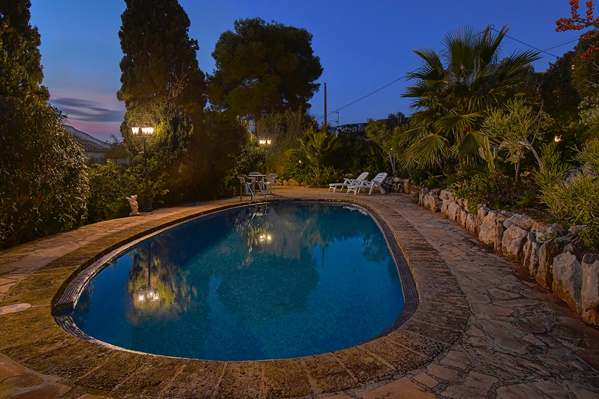 Piscina iluminada al anochecer rodeada de una terraza de piedra natural y un jardín mediterráneo maduro con palmeras y cipreses.