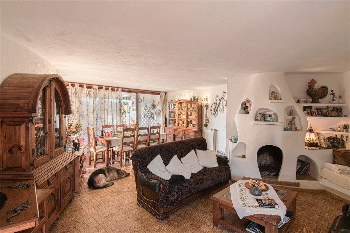 Salón comedor rústico con chimenea de obra esculpida, muebles de madera maciza y suelos de barro cocido. Grandes ventanales conectan con el exterior aportando luz natural.