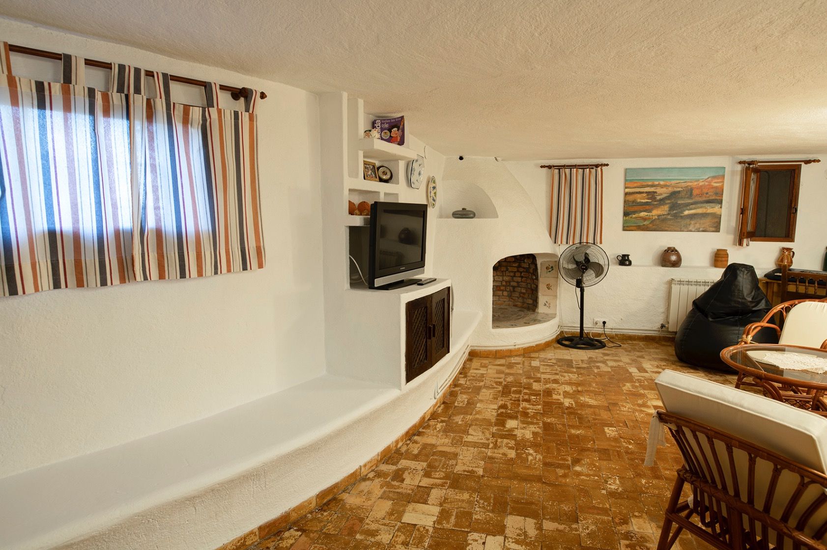 Salón de estilo rústico con chimenea de obra, suelos de barro tradicional y paredes blancas con estanterías y banco integrados.