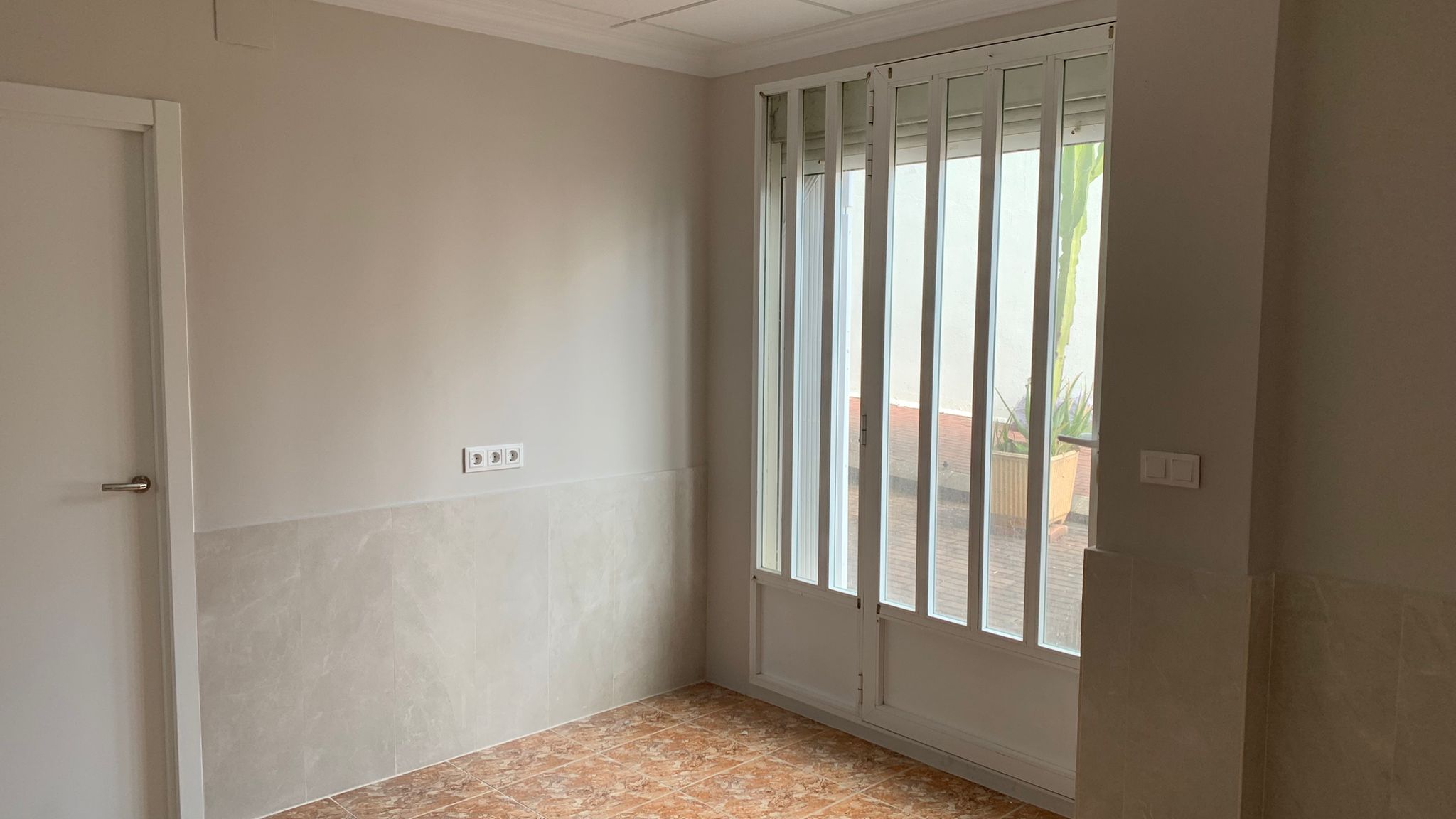 Habitación con suelos de gres, paredes parcialmente alicatadas y acceso directo al exterior mediante una puerta acristalada con rejas de seguridad blancas.