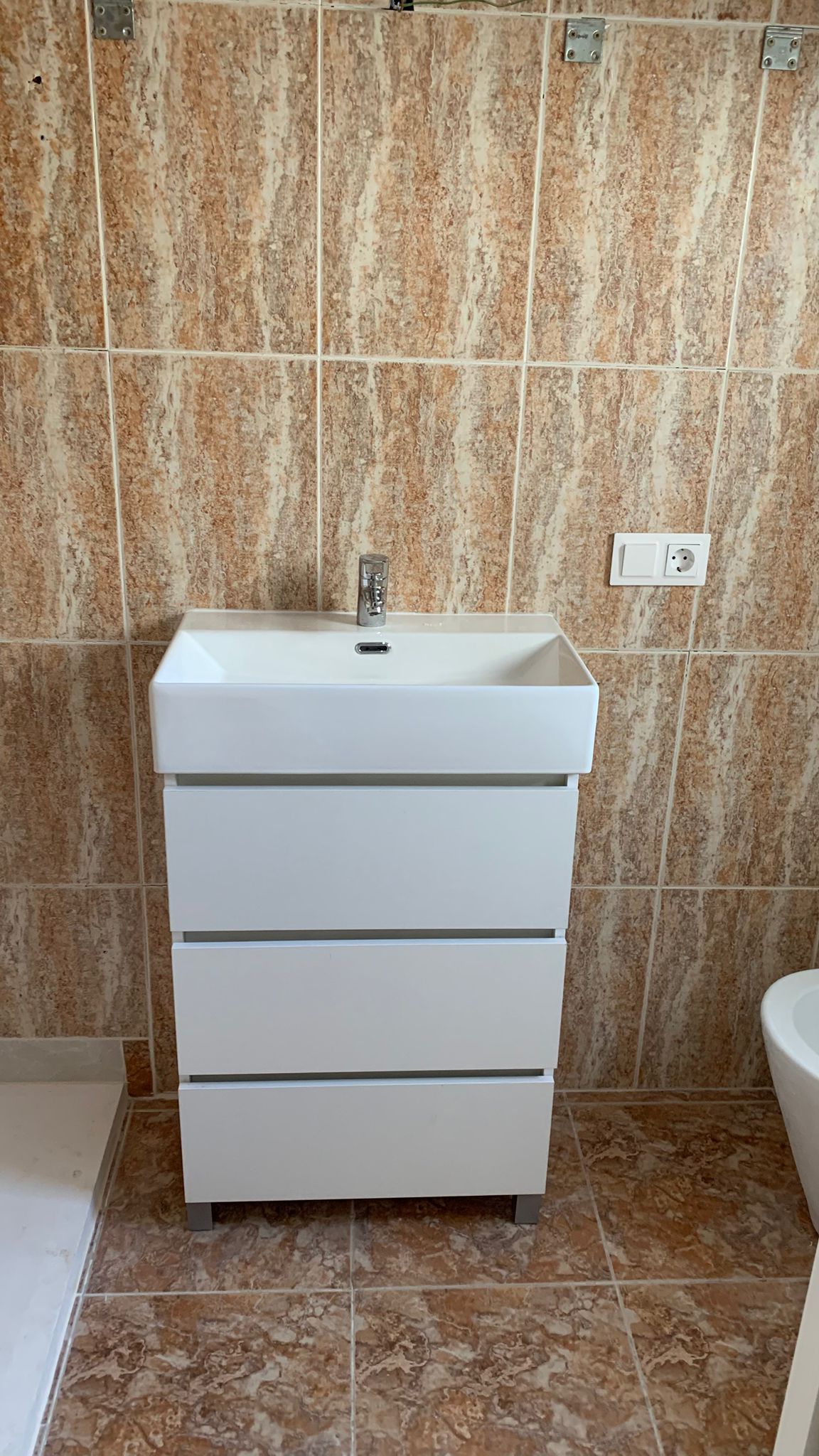 Mueble de baño moderno en color blanco con tres cajones y lavabo integrado, rodeado de azulejos cerámicos en tonos tierra y beige.