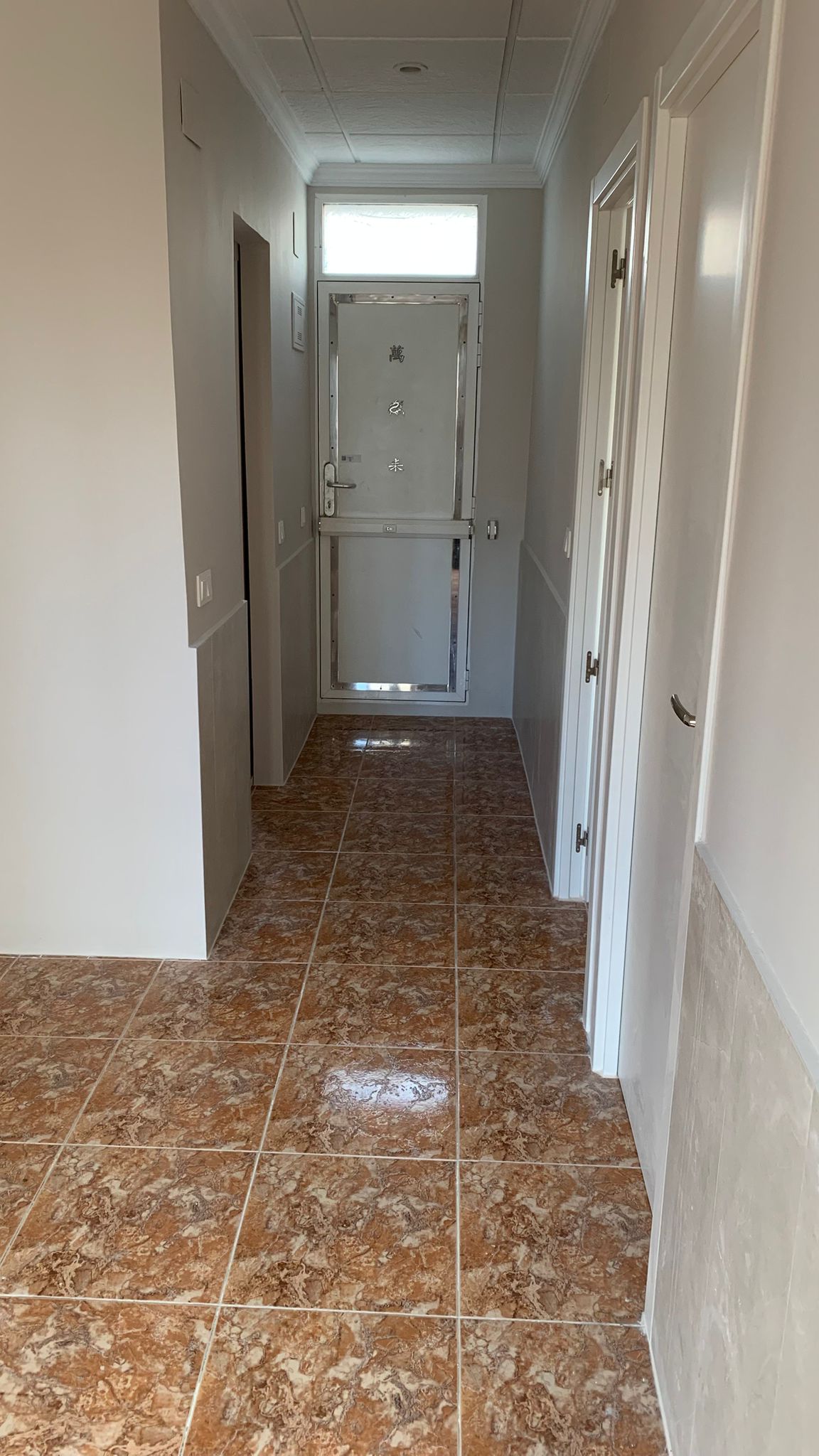 Pasillo de entrada con suelos de gres marmolado en tonos rojizos, paredes con zócalo cerámico gris y puerta de acceso reforzada con tragaluz superior.