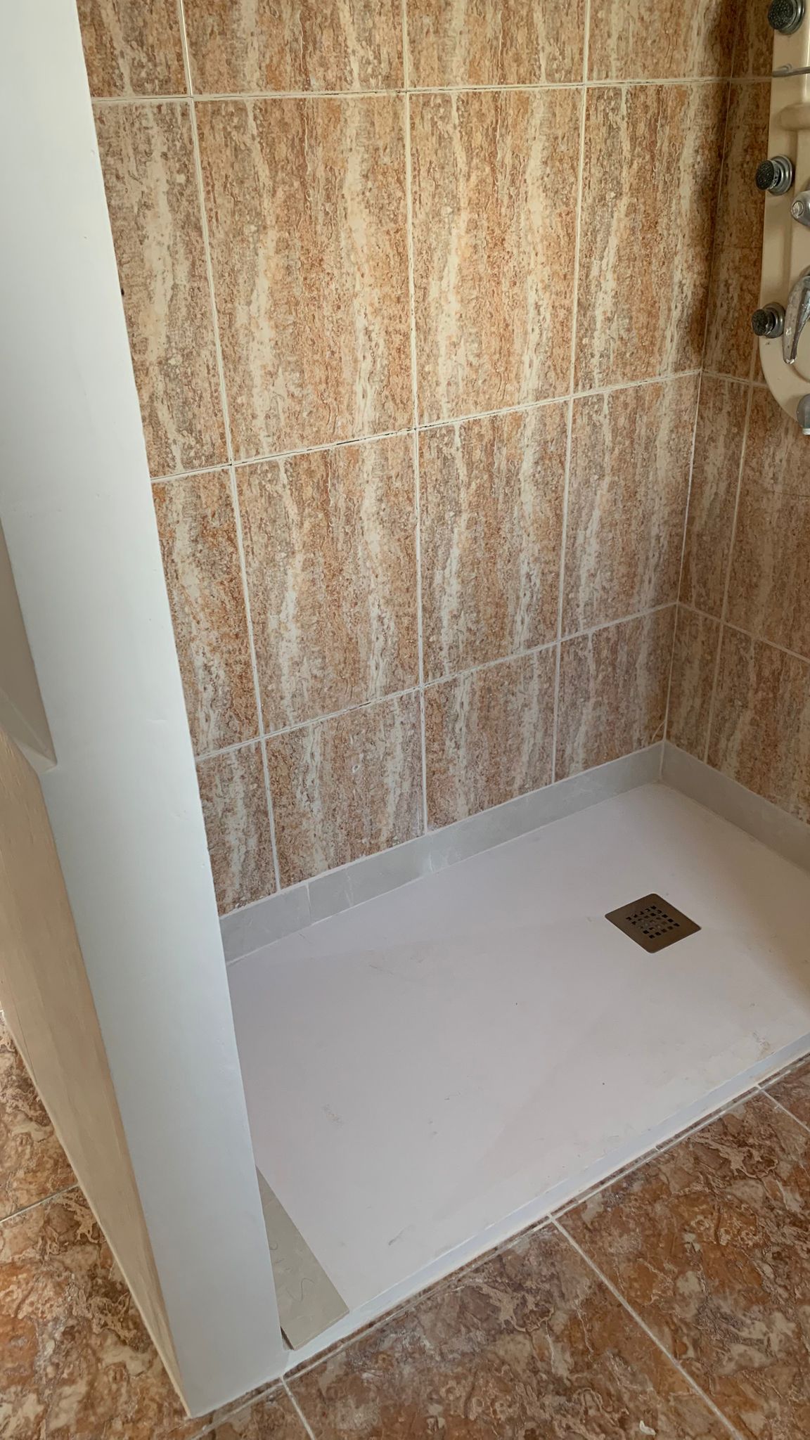 Plato de ducha blanco con mampara de obra, paredes revestidas en gres cerámico con efecto mármol y columna de hidromasaje integrada.
