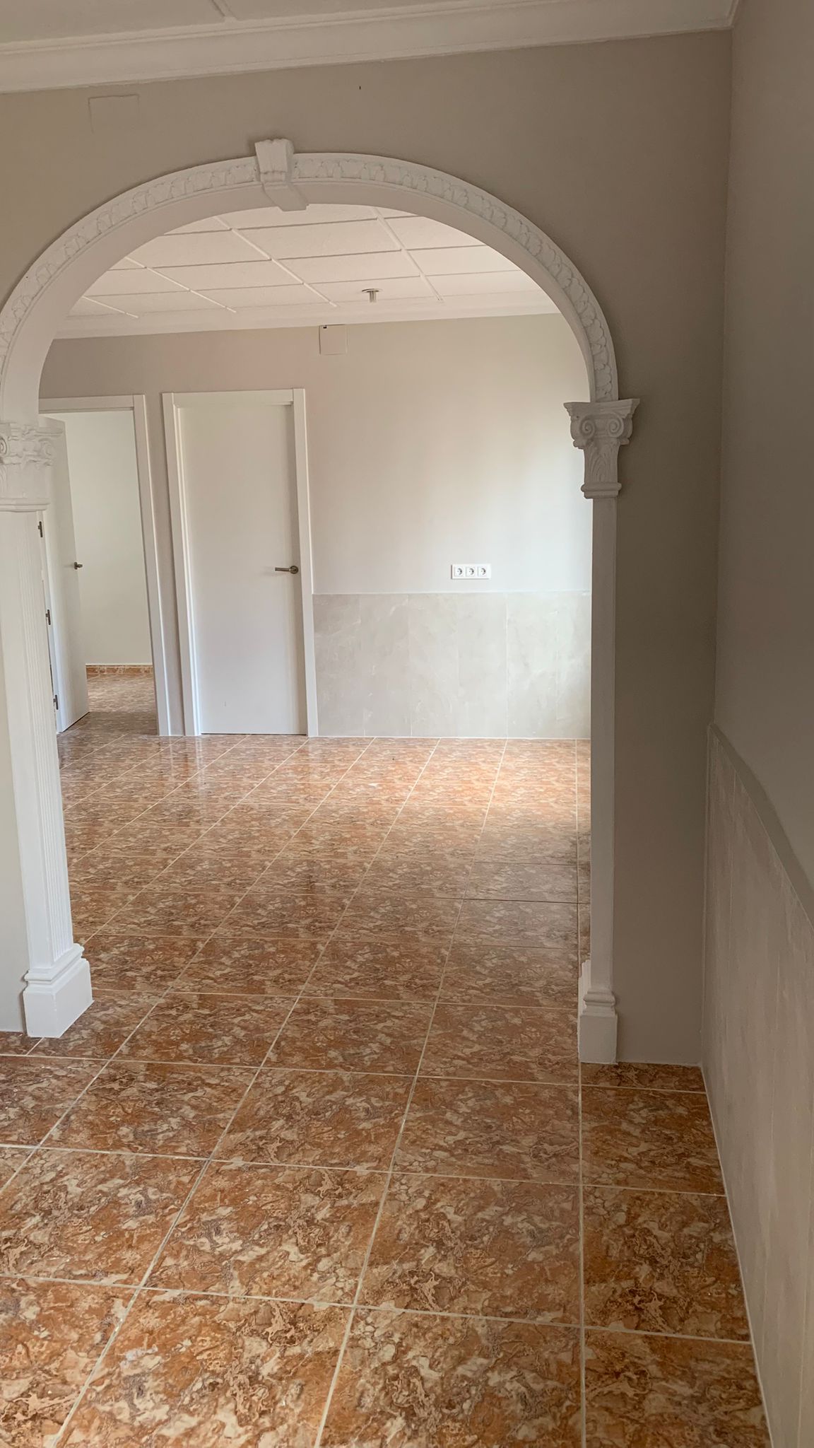 Vista a través de un arco decorativo blanco hacia una estancia con suelos de gres cerámico marrón, paredes en tonos neutros y puertas interiores blancas.
