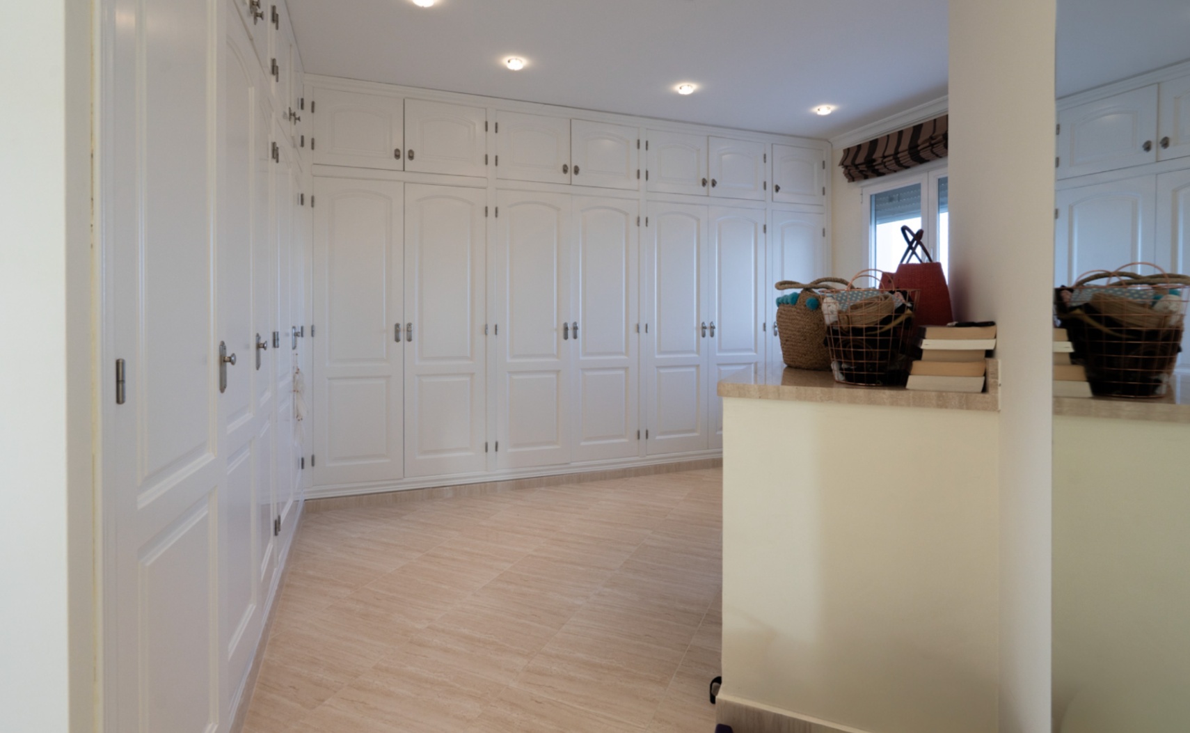 Amplio vestidor con armarios empotrados blancos de suelo a techo, molduras clásicas, iluminación integrada y suelos de gres porcelánico en tonos crema.