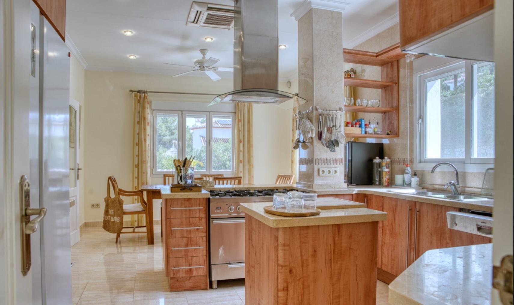 Cocina equipada con isla central, encimeras de piedra y electrodomésticos de acero inoxidable. Destaca la campana extractora de diseño, los suelos cerámicos y la zona de comedor.